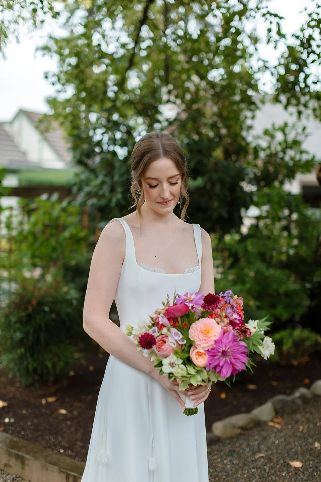 A woman in a white dress holding a colorful bouquet of flowers outdoors with trees and houses in the background.
