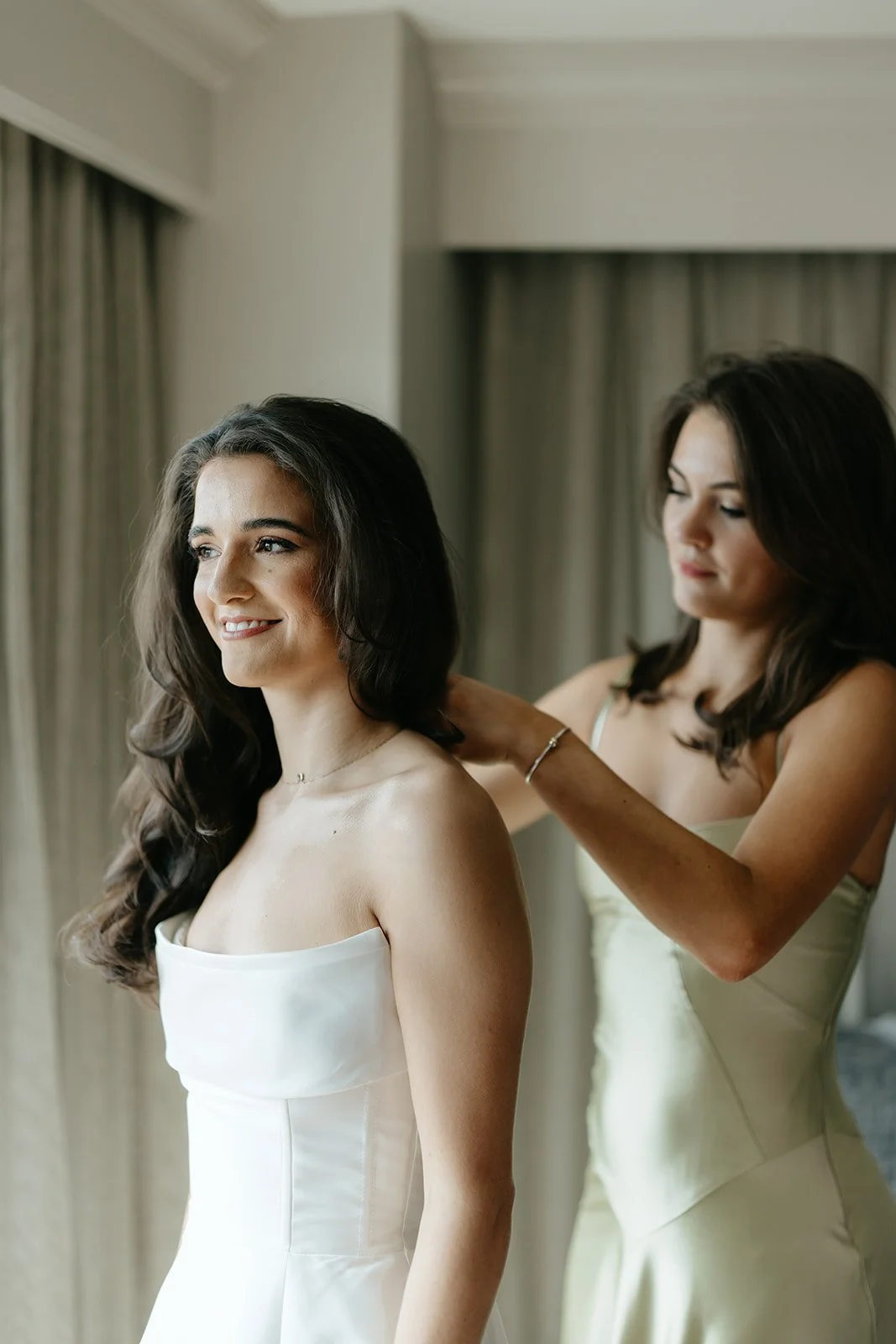 A bride in a white strapless wedding gown smiling while a woman, possibly her Maid of Honor, assists her with the dress.