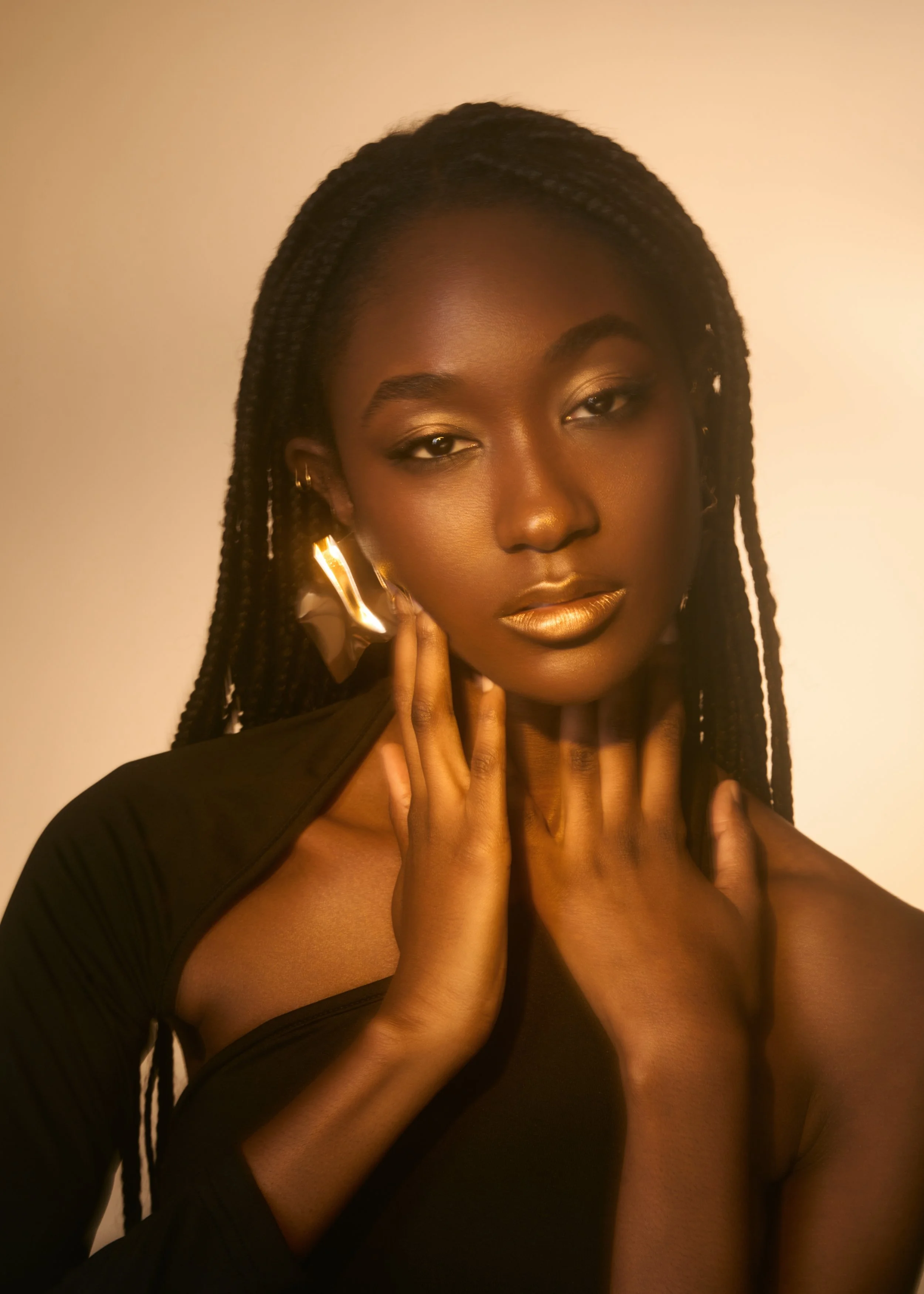 Close-up of a woman with braided hair, wearing gold lipstick and earrings, touching her face with her hands, in a black outfit, against a plain beige background.