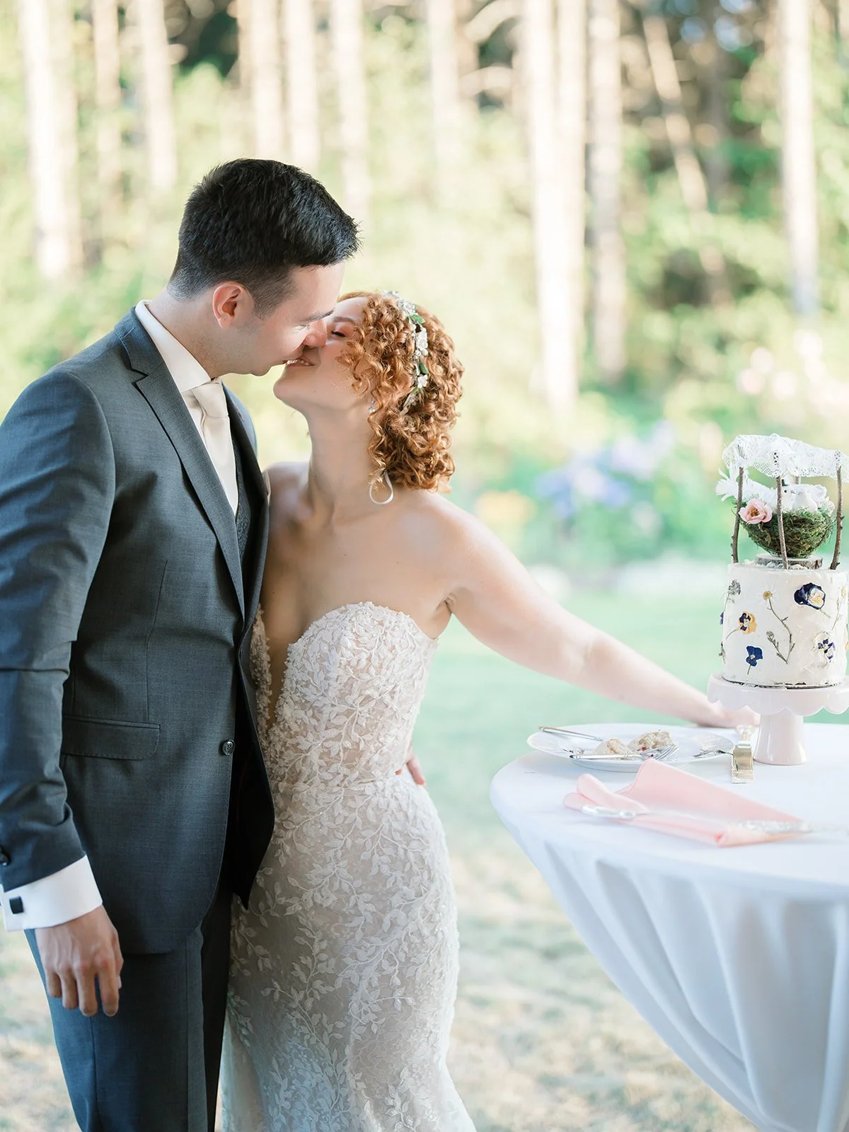 A bride and groom kiss at their outdoor wedding reception, with a wedding cake on a table nearby.