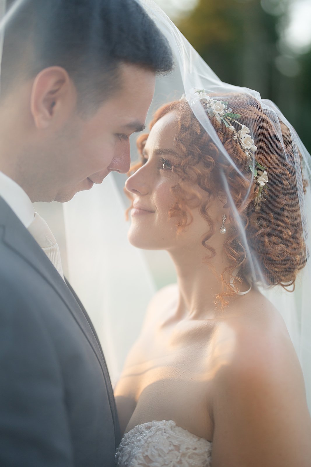 Bride and groom face-to-face, smiling with a veil over their heads, outdoors with soft natural light.