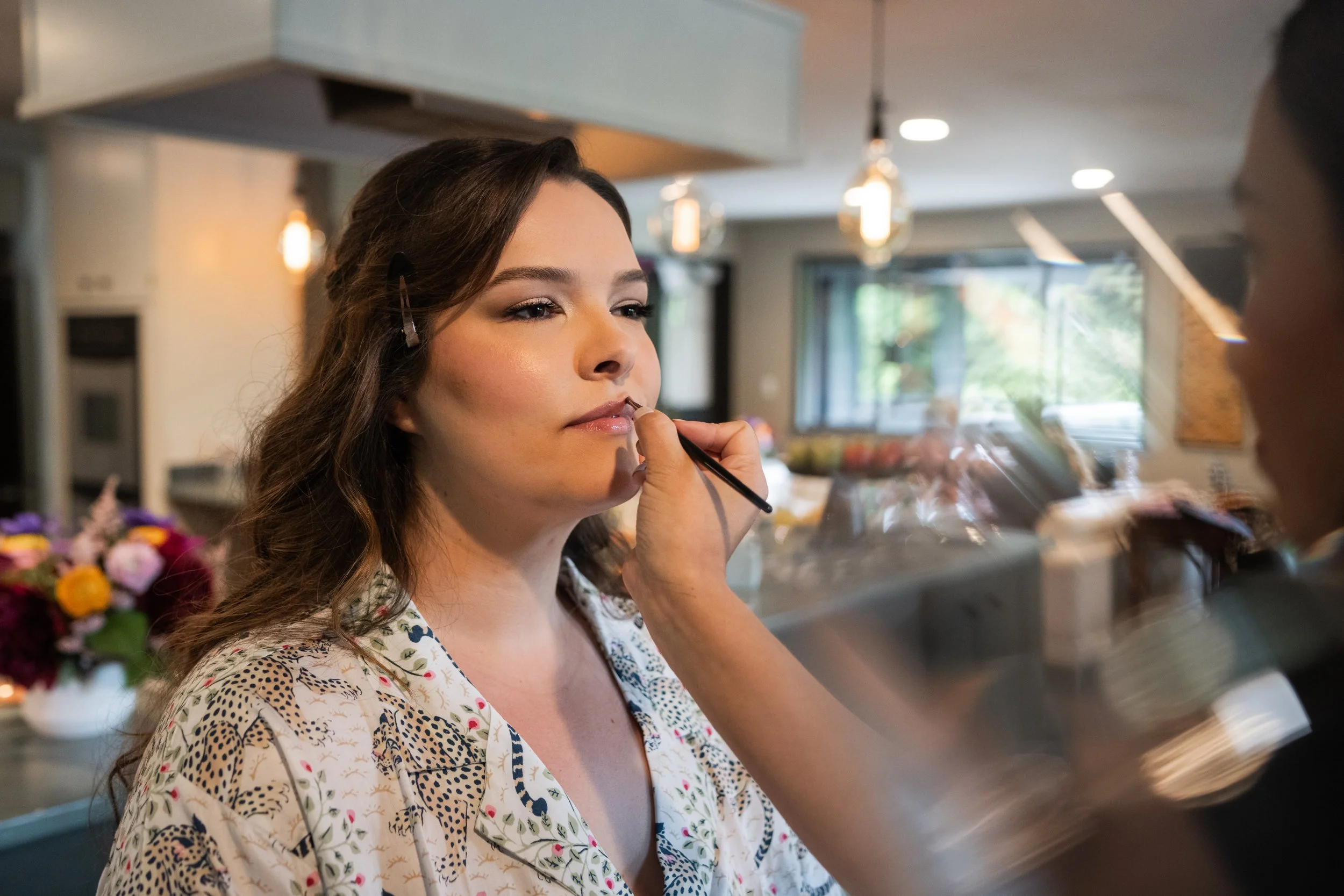 A woman with brown wavy hair having makeup applied by a makeup artist in a modern kitchen with pendant lights, large windows, and floral decor.