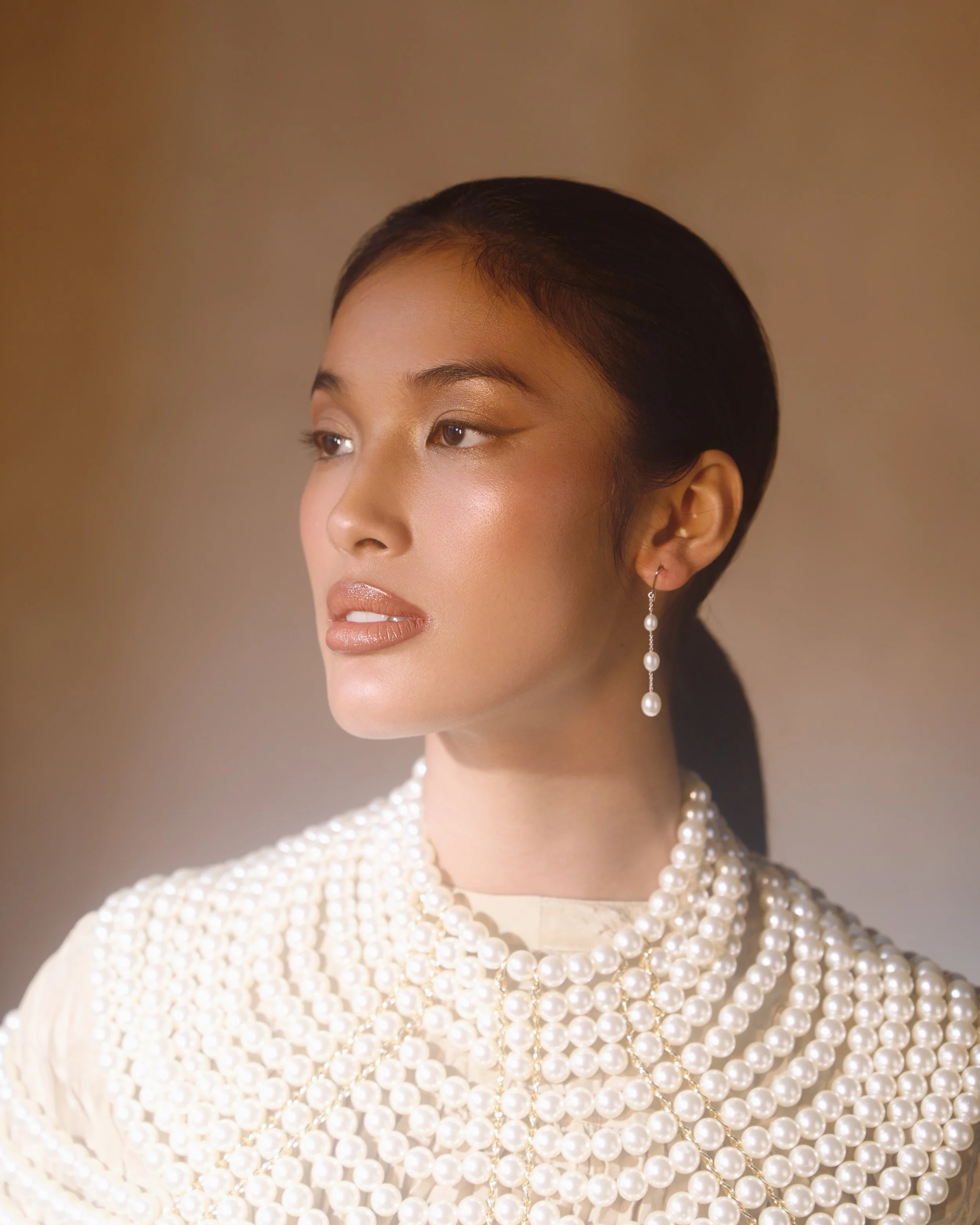 A woman with dark hair styled in a sleek bun, wearing pearl jewelry including earrings, a necklace, and a pearl-embellished dress, posed against a neutral background.