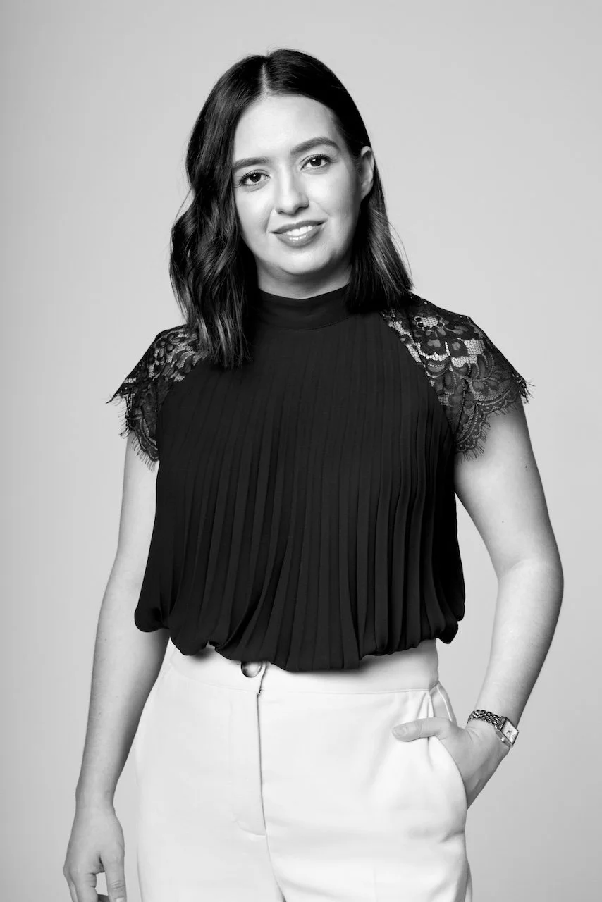 Black and white portrait of a woman with shoulder-length dark hair, wearing a black pleated top with lace details on the shoulders, white pants, and a watch on her left wrist, standing against a light grey background.
