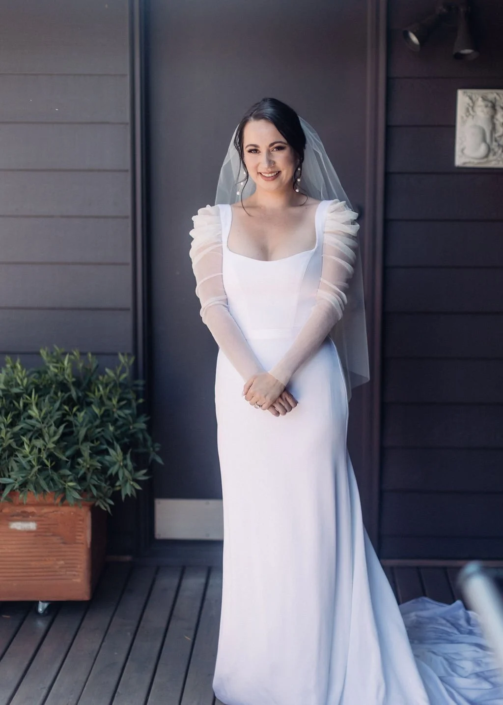 A woman in a white wedding dress with sheer puff sleeves and a veil, standing outdoors against a dark wooden wall, smiling and holding her hands together.
