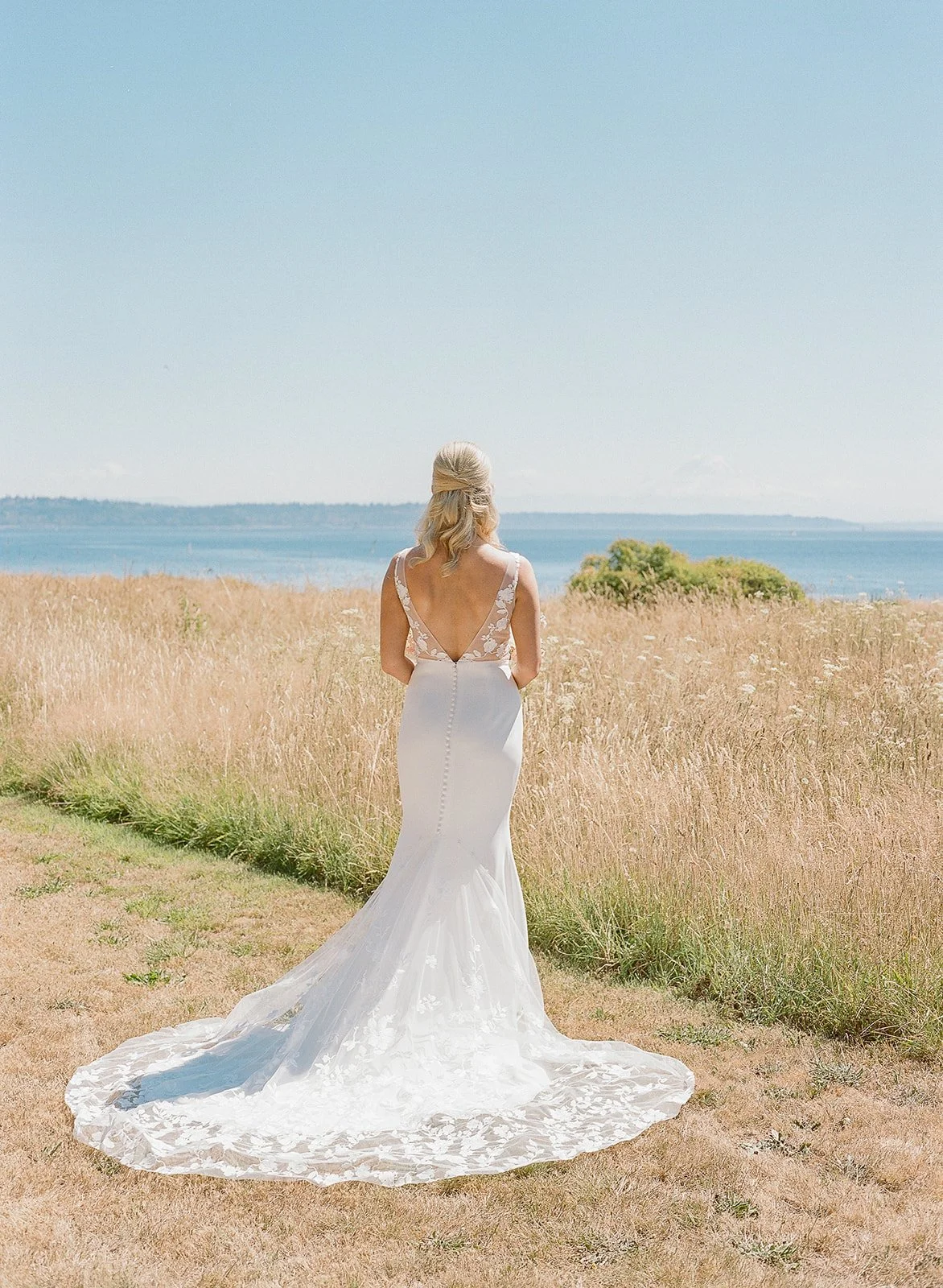 A woman in a white wedding dress with lace details, standing on a grassy area near a sandy path, looking out over a field of tall, golden grass and a body of water under a clear blue sky.