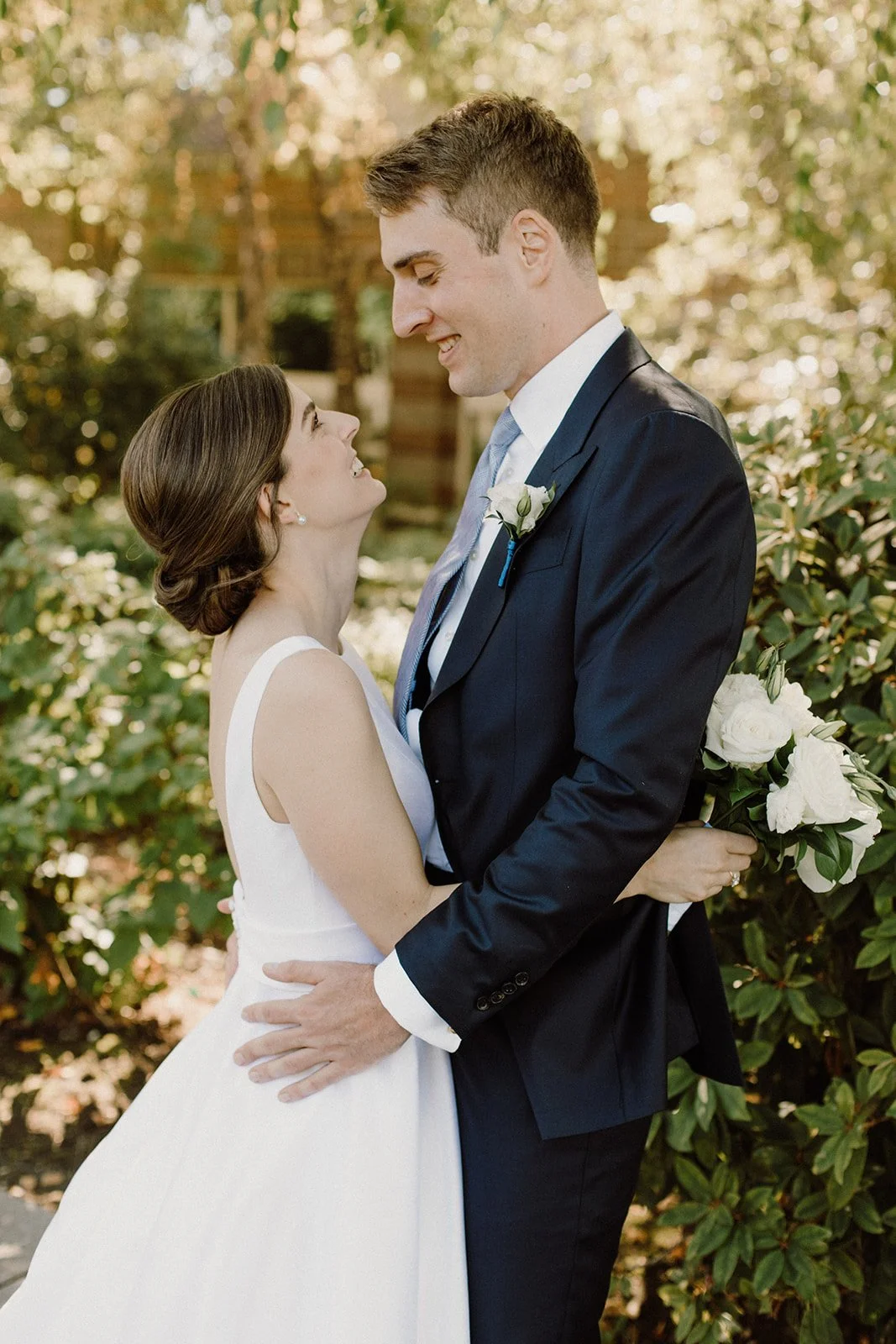 A bride and groom looking into each other's eyes outdoors, with greenery and sunlight in the background. The bride is in a white wedding dress and the groom in a dark suit with a boutonniere and a bouquet of white flowers.