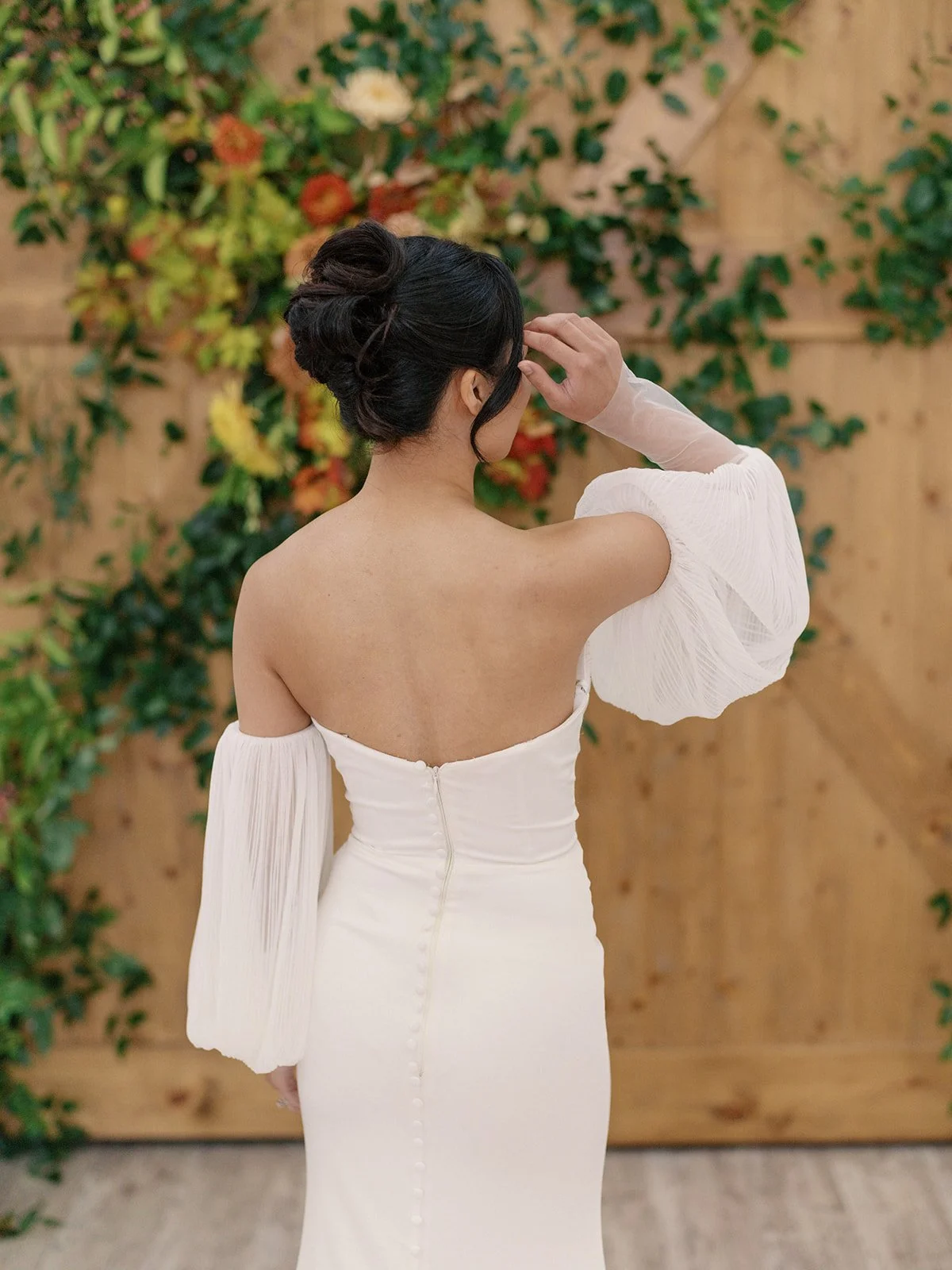 A woman in a white off-the-shoulder dress with puffed sleeves and a zipper back, standing with her back to the camera, touching her hair. She is in front of a wooden wall decorated with green leaves and flowers.