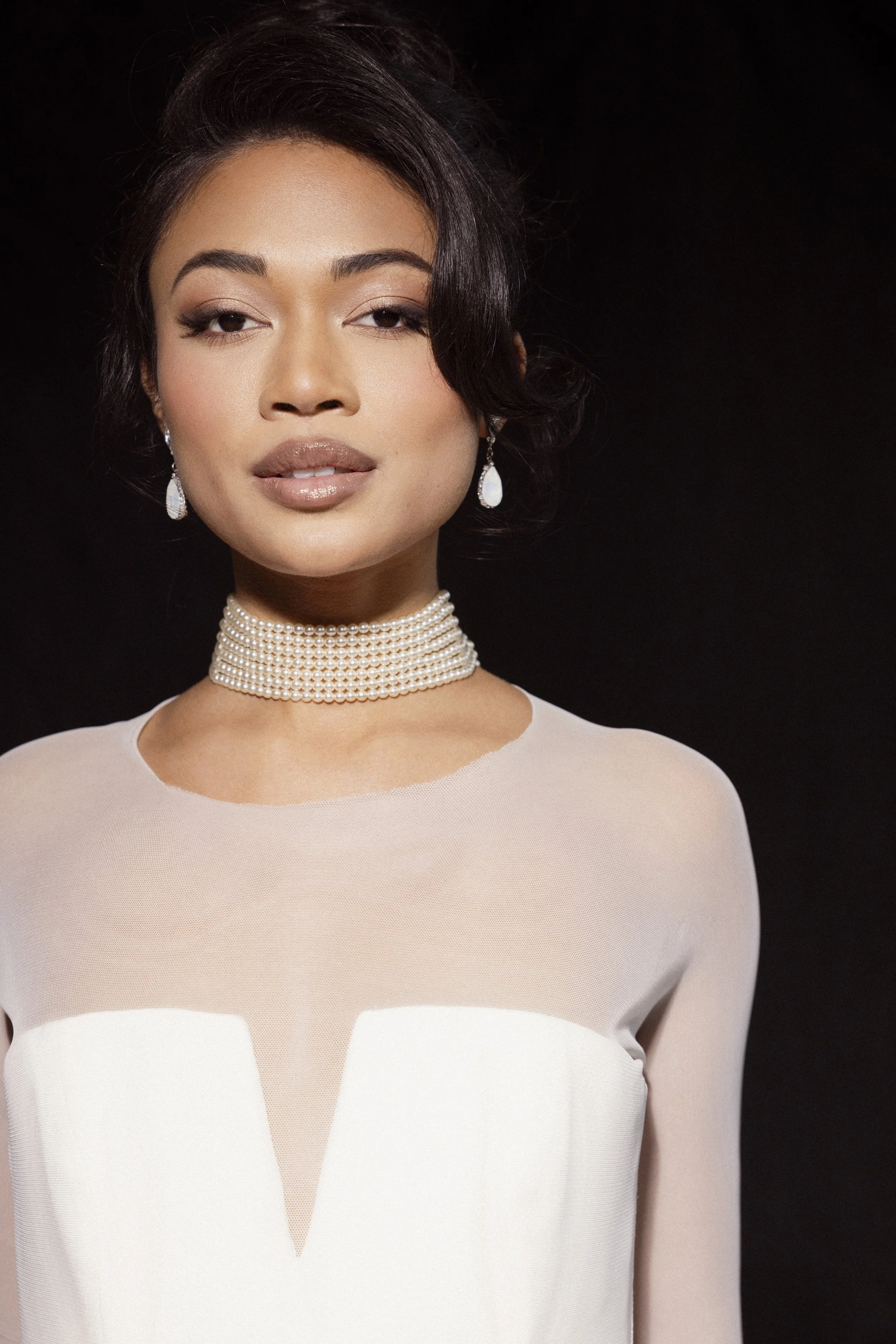 A woman with dark hair styled in soft waves, wearing elegant pearl earrings and a multi-strand pearl choker, dressed in a white dress with a sheer, cut-out design, posing against a black background.