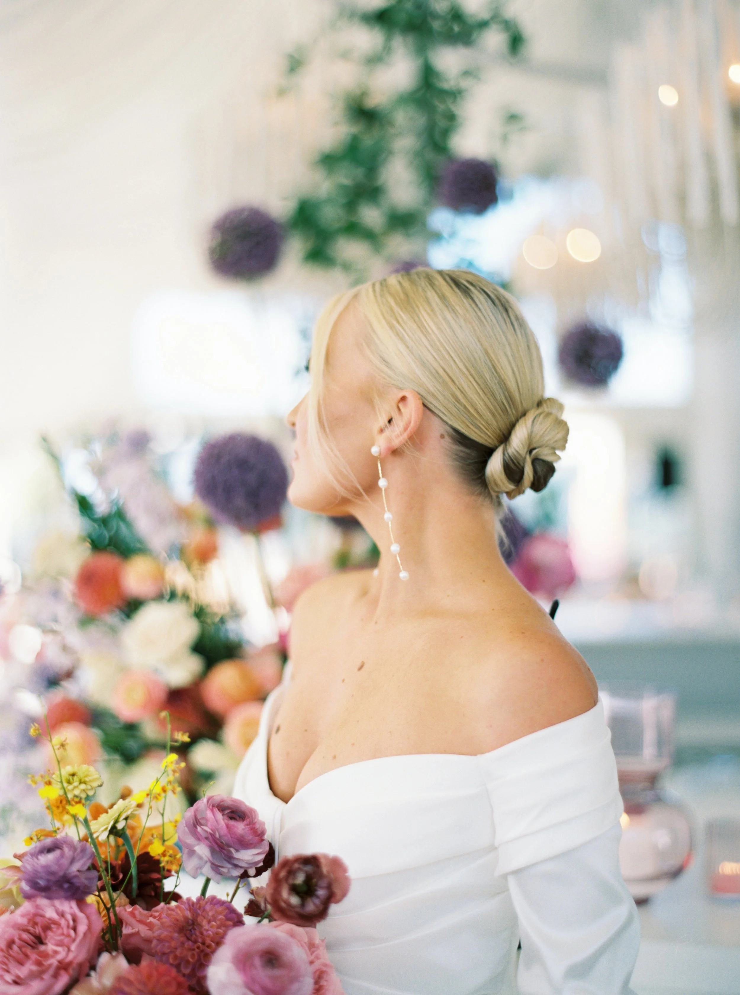 A woman in an off-shoulder white dress with blonde hair styled in a low bun, standing in front of a colorful flower arrangement, with greenery and blurred lights in the background.