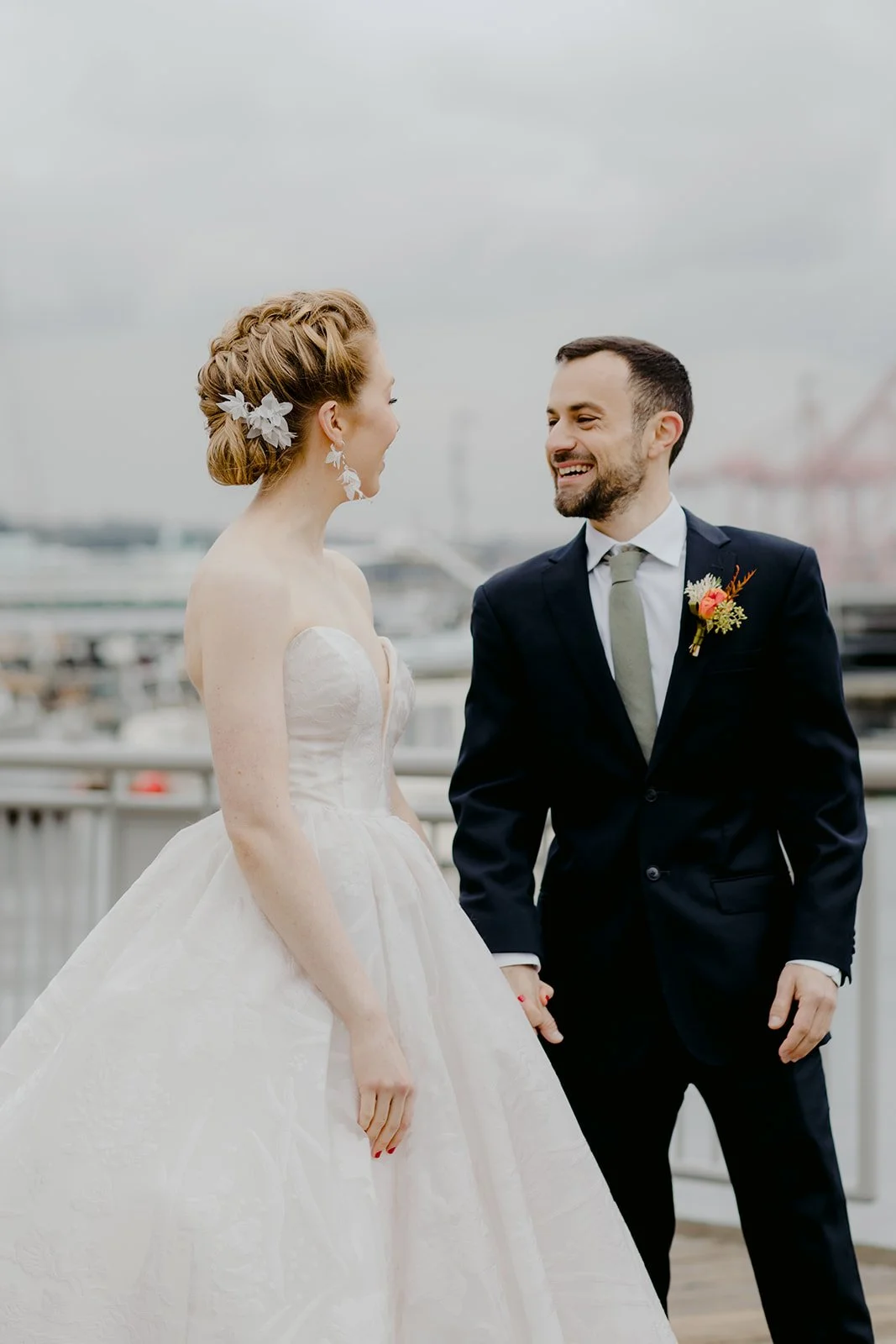 A bride and groom holding hands on a cloudy day, smiling at each other, with a waterfront and boats in the background.