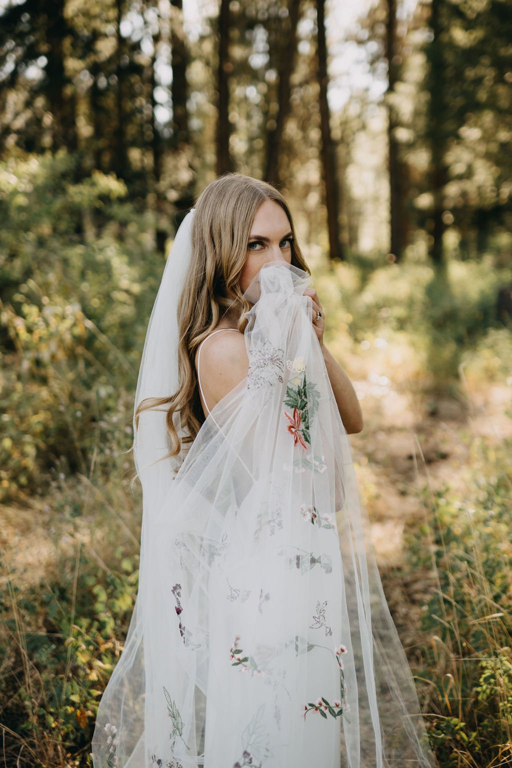 A bride with long wavy hair wearing a white dress and veil, standing in a forested area, partially covering her face with a sheer embroidered veil.