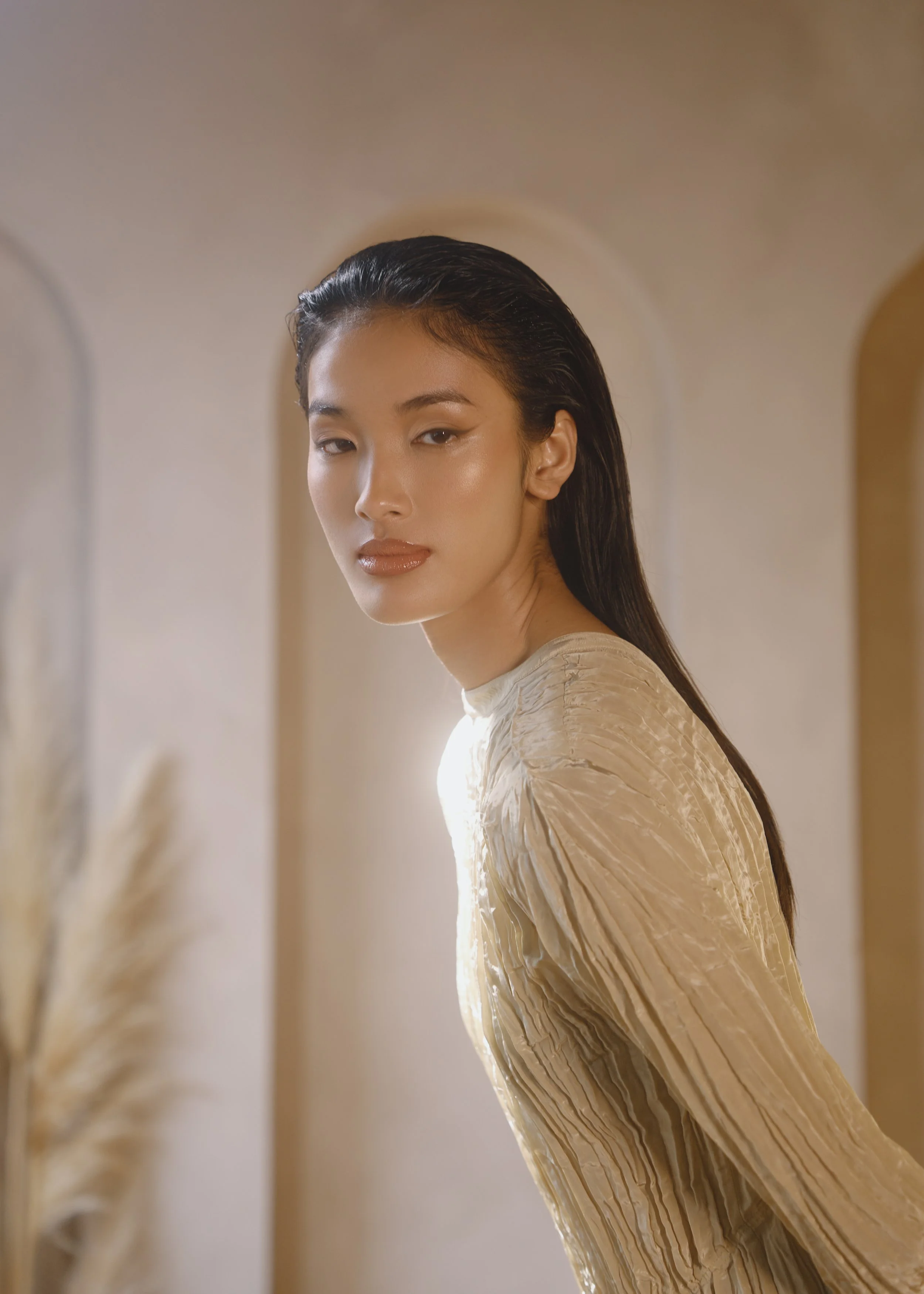 A woman with wet, slicked-back hair looking at the camera, standing in a neutral-colored room, wearing a textured beige long-sleeve top.