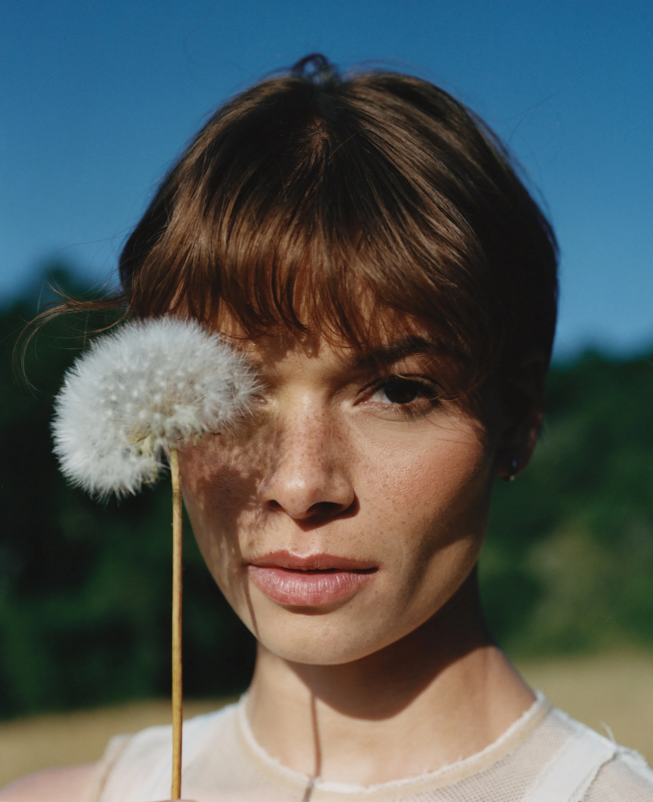 A young woman with short brown hair and freckles holding a dandelion flower close to her face outdoors on a sunny day.