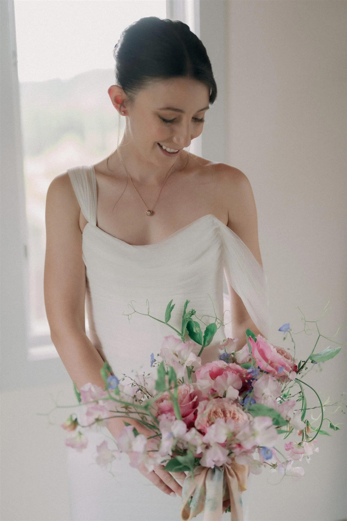 A woman in a white dress holding a large bouquet of pink and purple flowers, smiling while looking down.