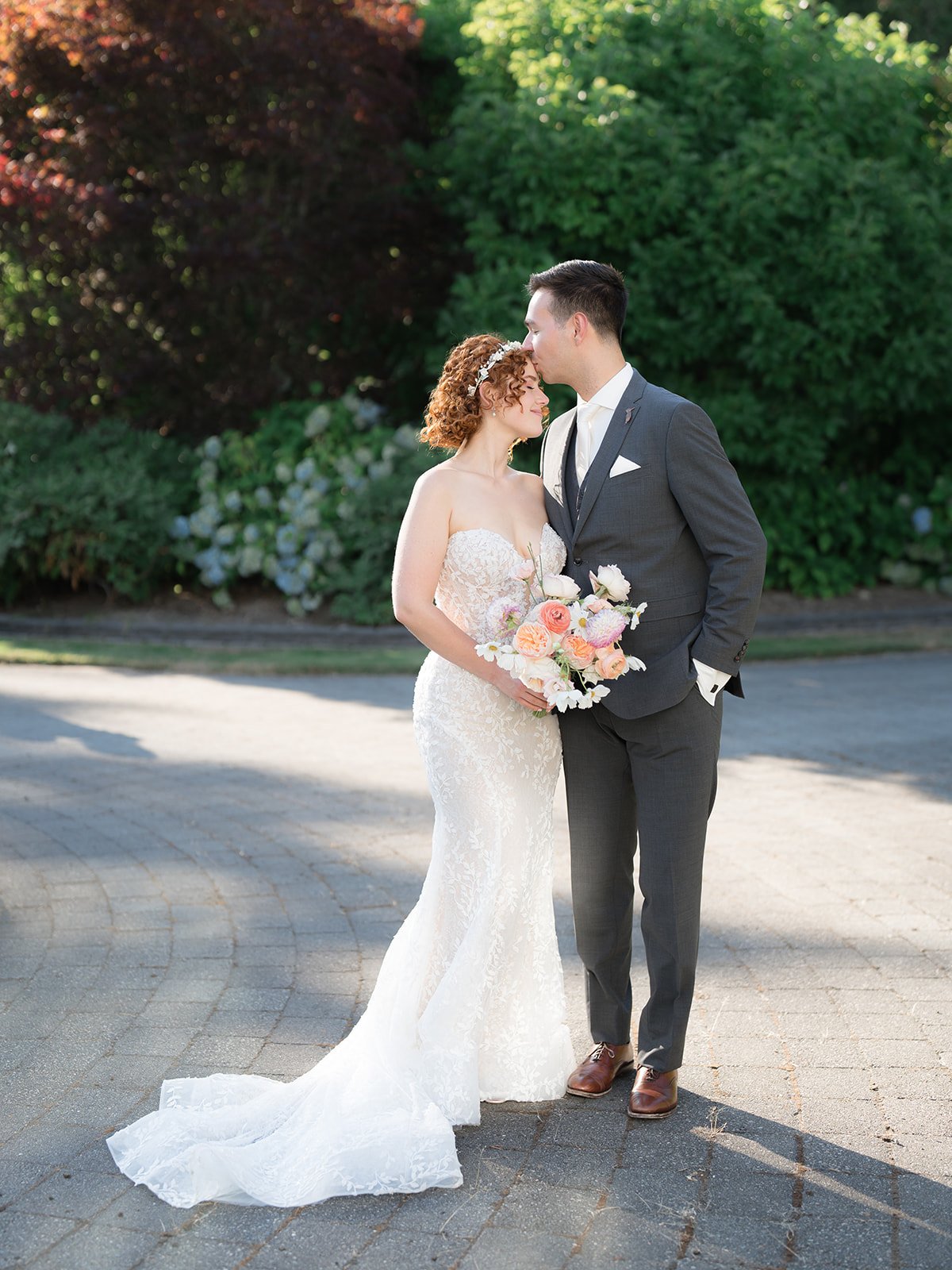 A bride in a white lace wedding dress and a groom in a gray suit share a tender moment outdoors. The bride holds a colorful bouquet, and the groom gently kisses her forehead. Bright sunlight and lush green foliage create a romantic atmosphere.