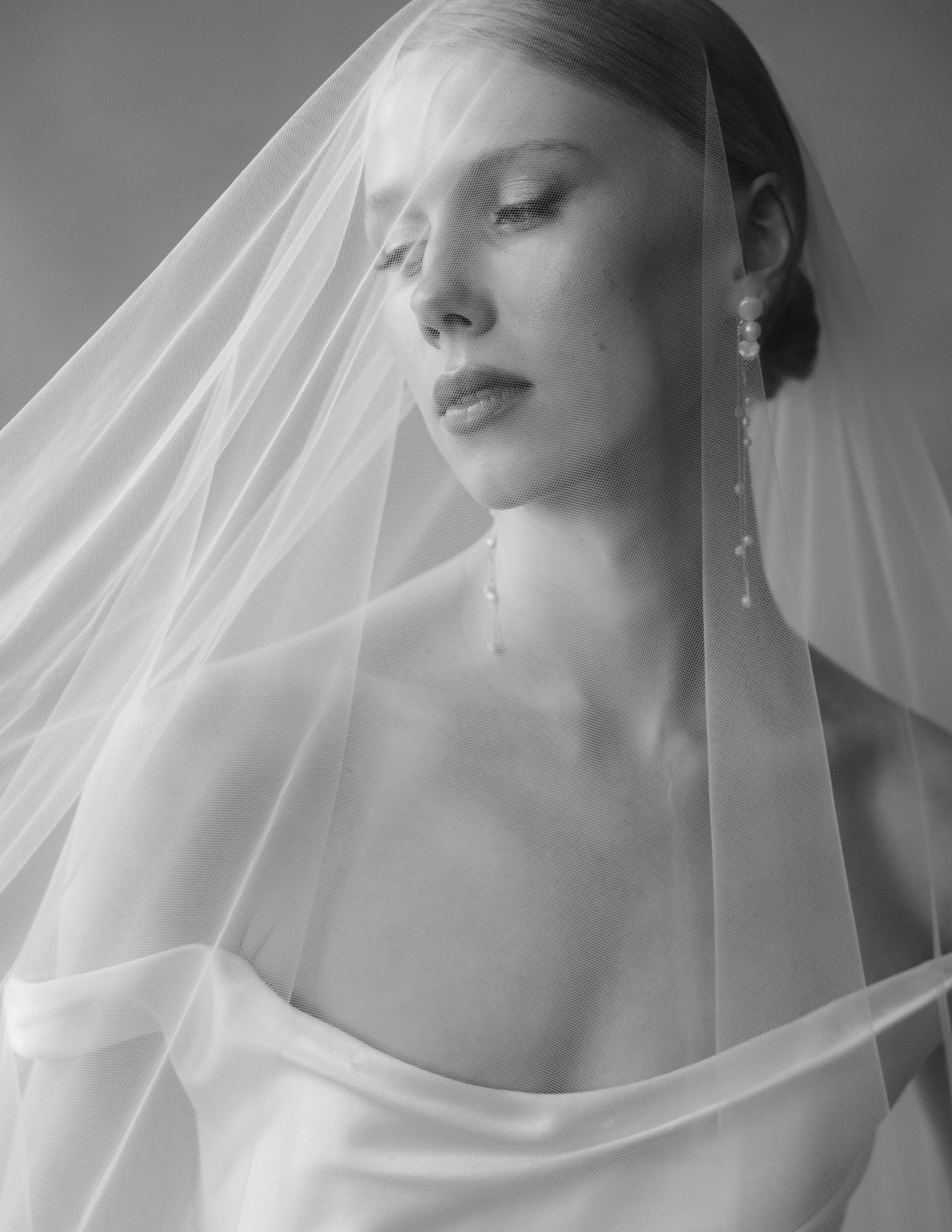 A close-up black and white portrait of a bride with her eyes closed, wearing pearl earrings and a veil.