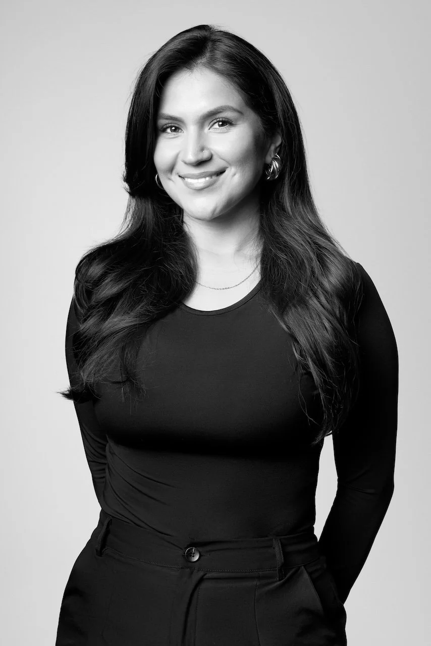 Woman with long wavy dark hair smiling, wearing hoop earrings, a black top, and pants, posed with one hand in pocket against a plain background.