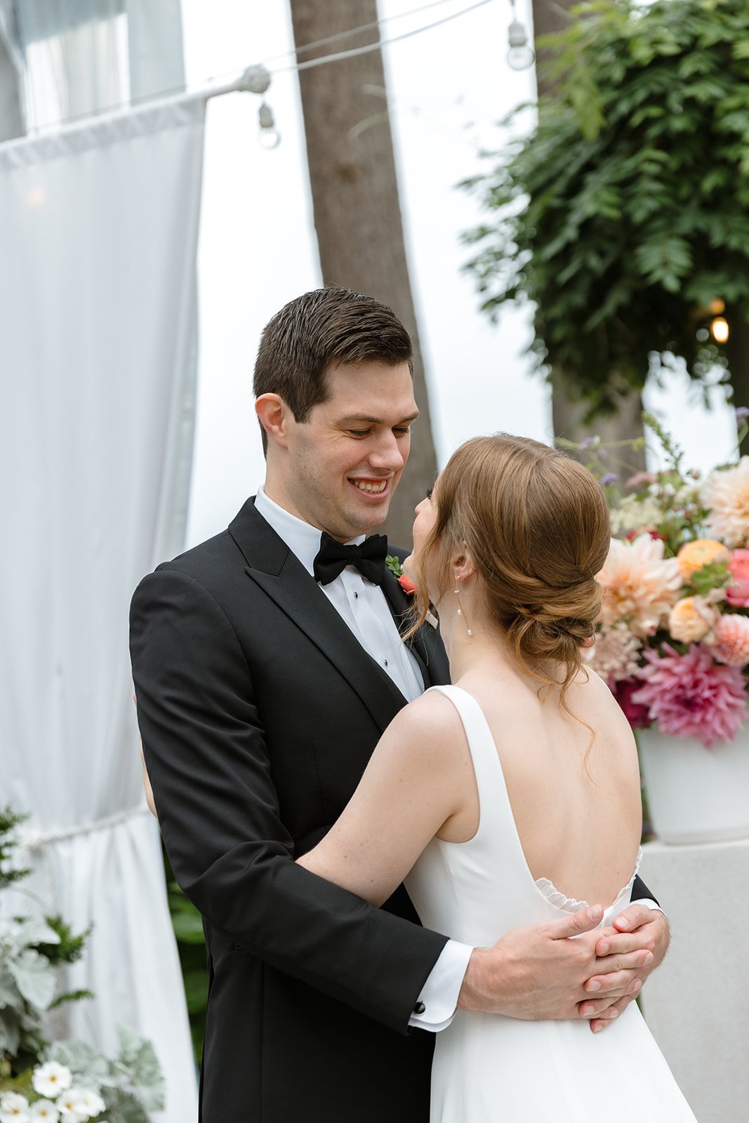 A wedding couple dancing outdoors, the groom in a black tuxedo and the bride in a white dress, smiling at each other with flowers and greenery in the background.