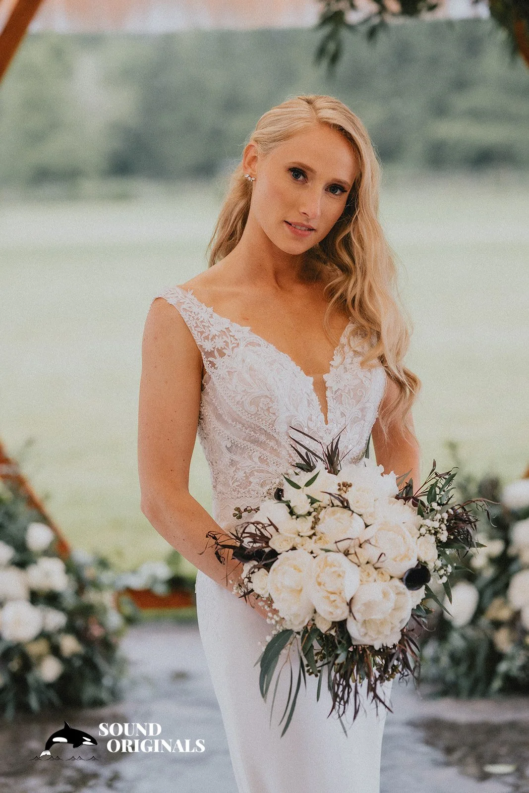 A bride in a white lace wedding gown holding a bouquet of white flowers and greenery, standing outdoors with a blurred green background.