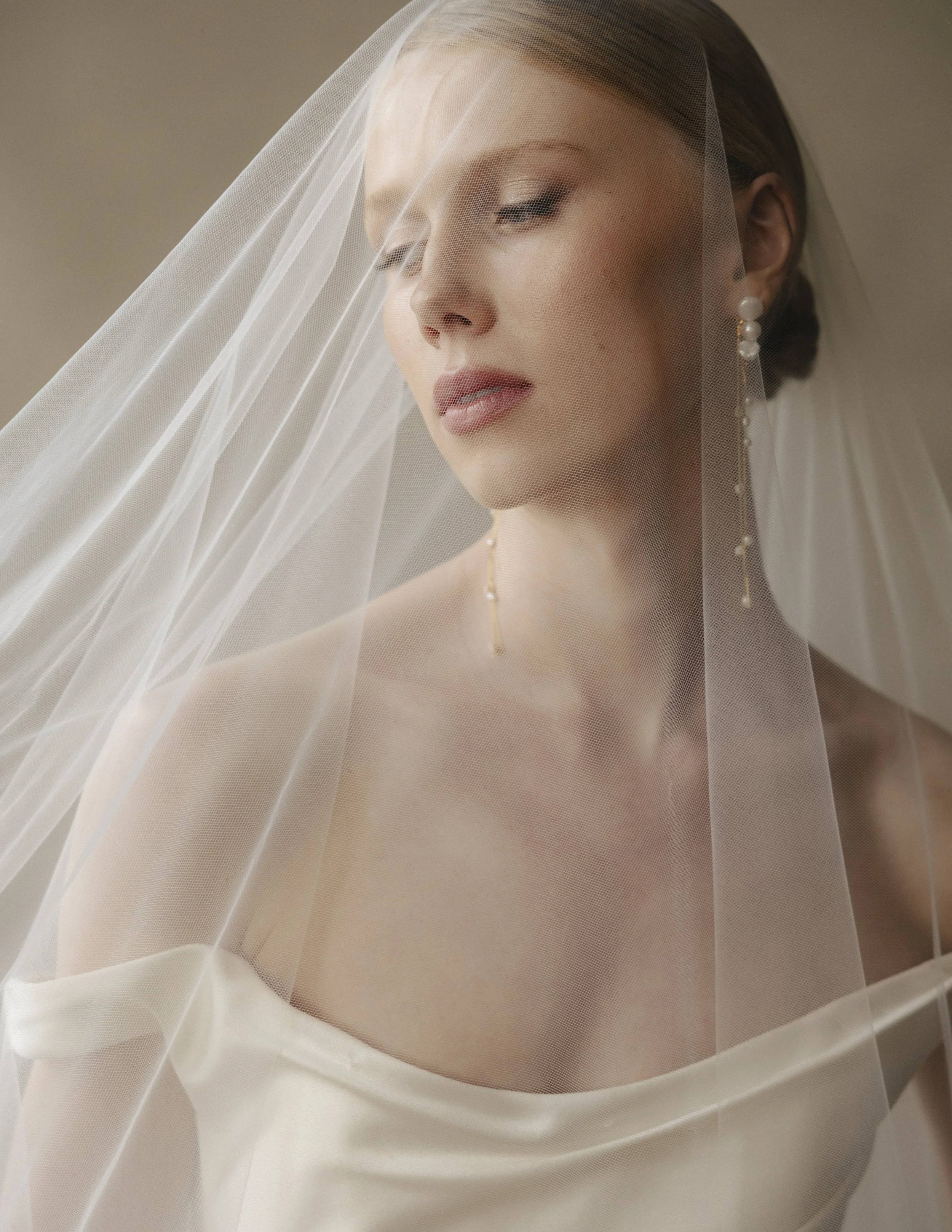A close-up of a woman with makeup, pearl earrings, wearing a veil, and a strapless top or dress, with soft lighting and a neutral background.