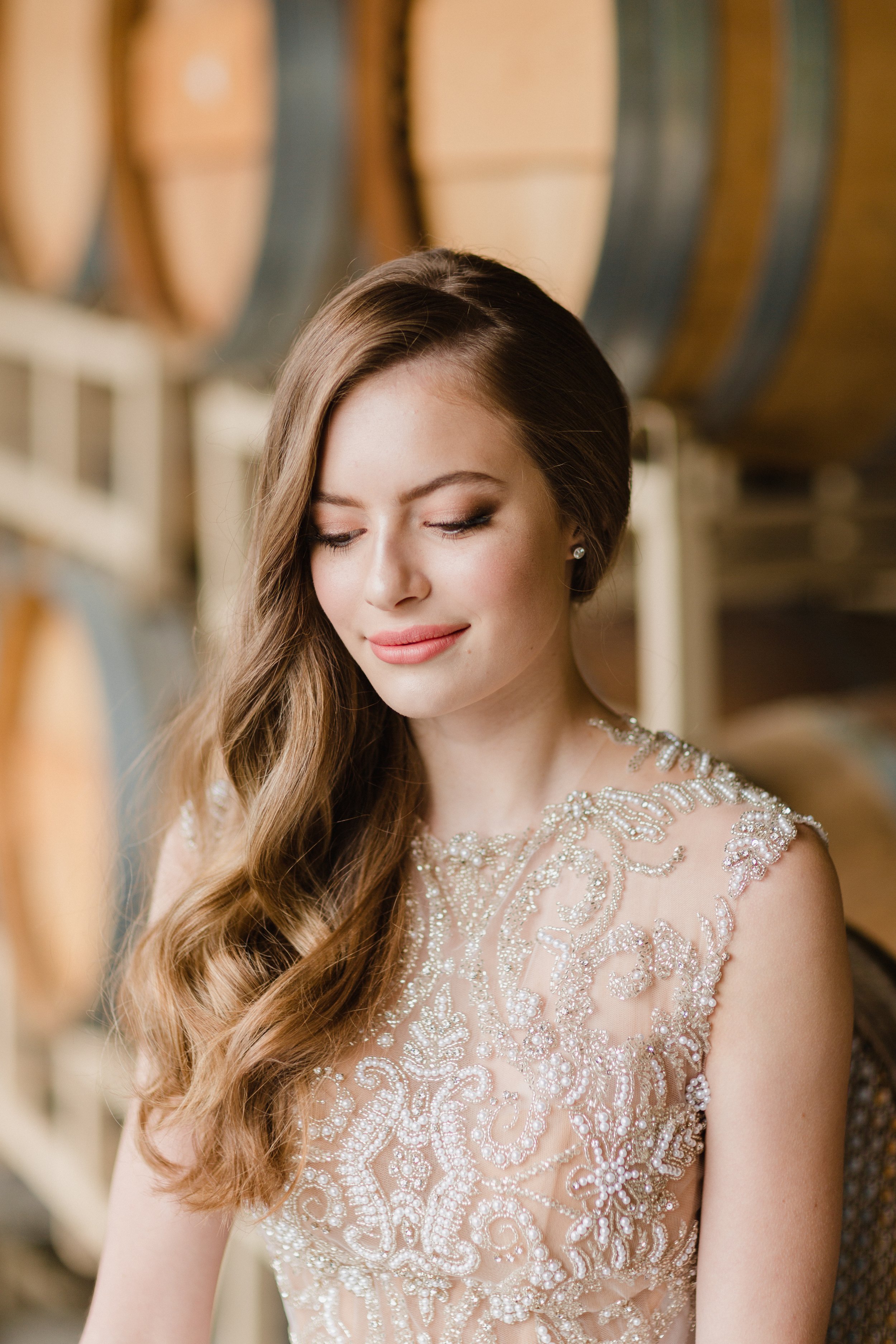 A young woman with long, wavy brown hair and light makeup, wearing a beaded, embellished dress, standing in front of rows of wooden chairs.
