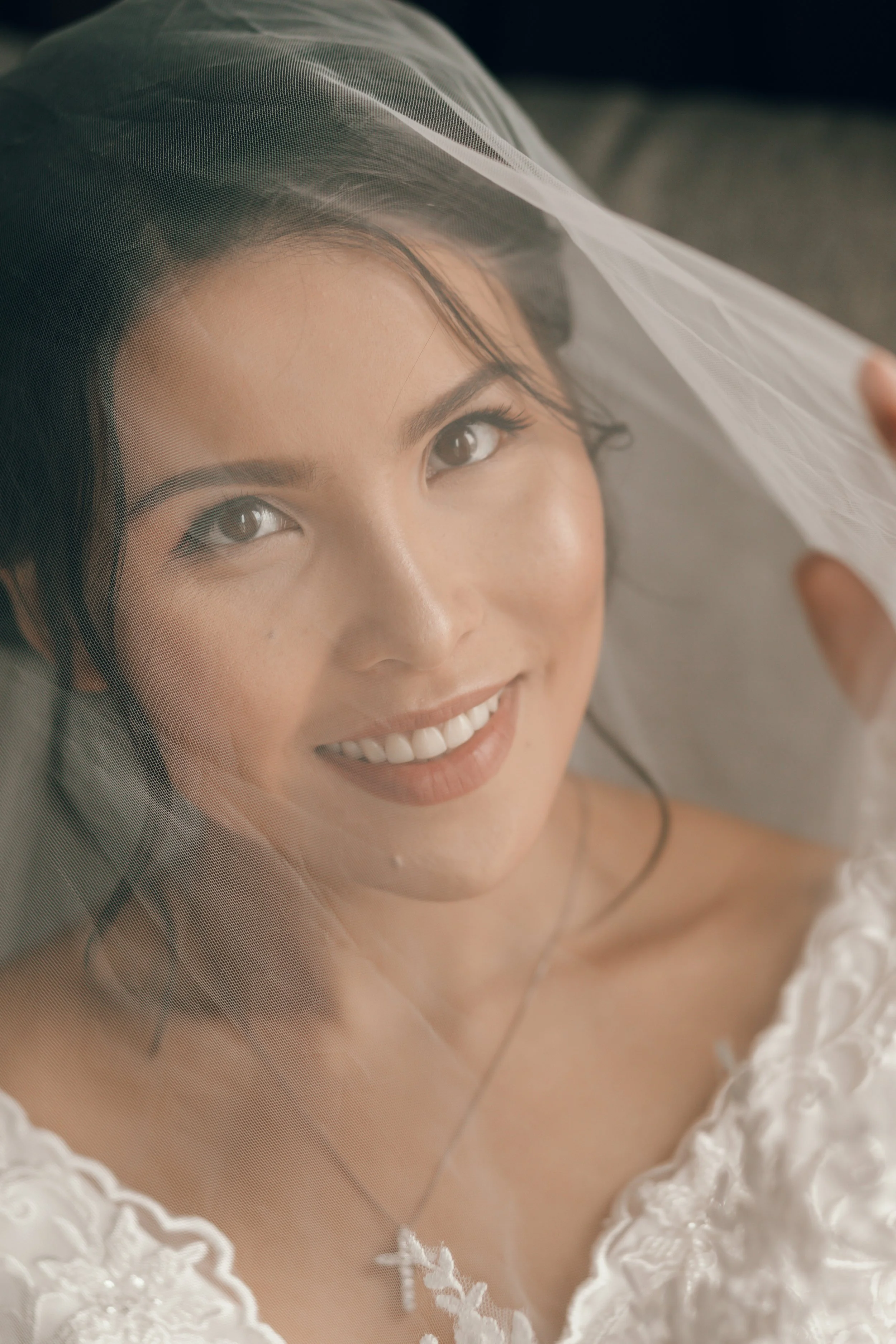 A smiling woman with dark hair and brown eyes, lying down and holding a sheer veil over her face, wearing a white lace dress.