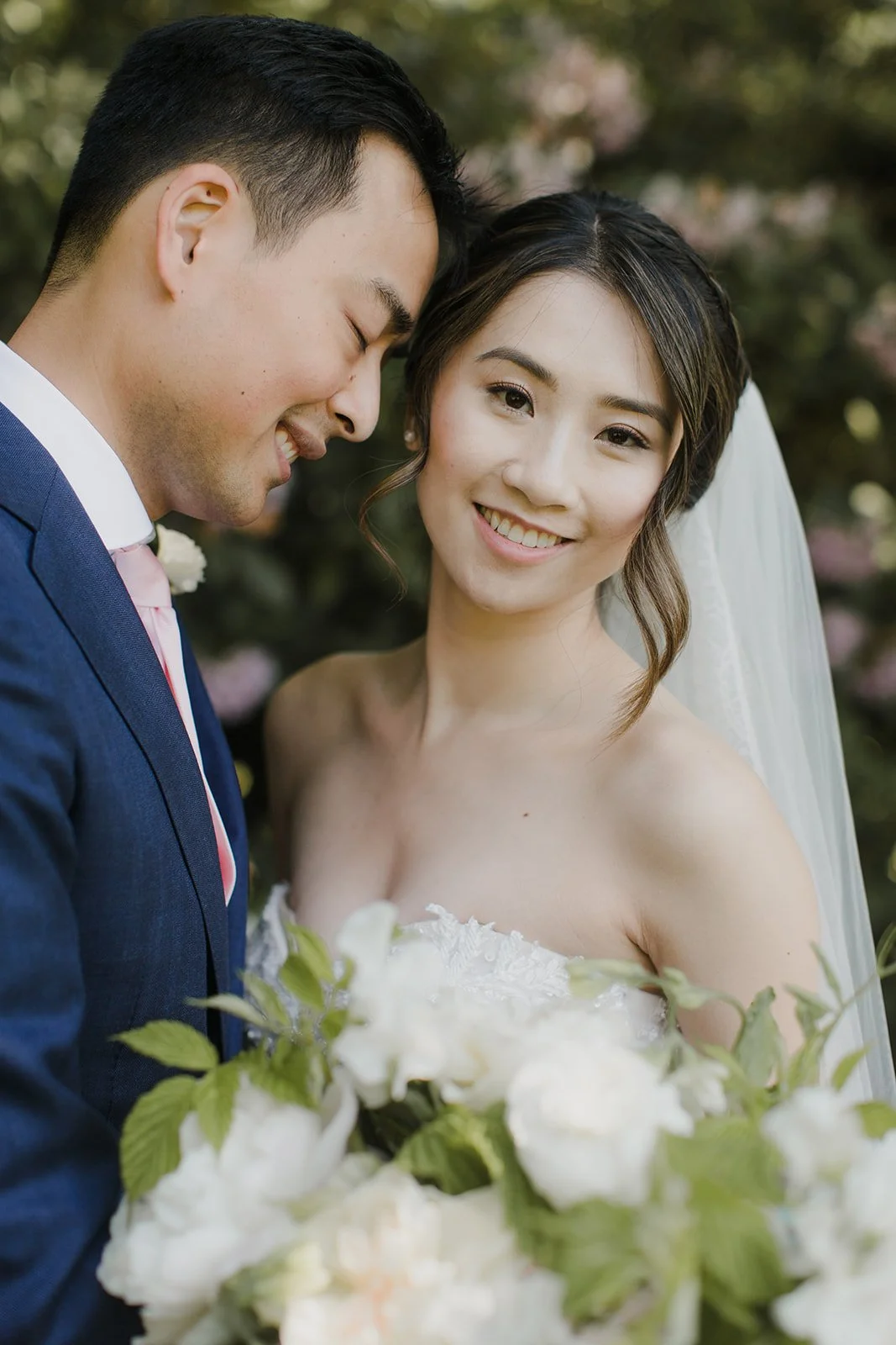 A newlywed couple smiling outdoors, with the groom in a navy suit and the bride holding a bouquet, surrounded by greenery.