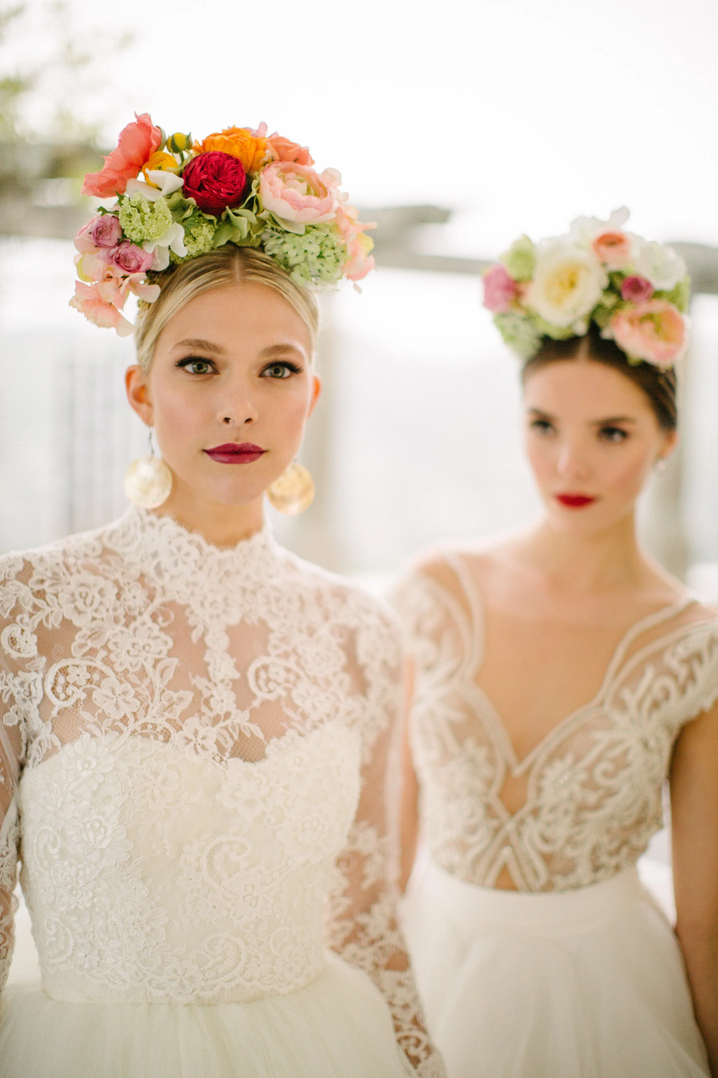 Two women dressed in white lace wedding gowns with floral headpieces, standing indoors with white walls, one facing the camera and the other slightly turned.