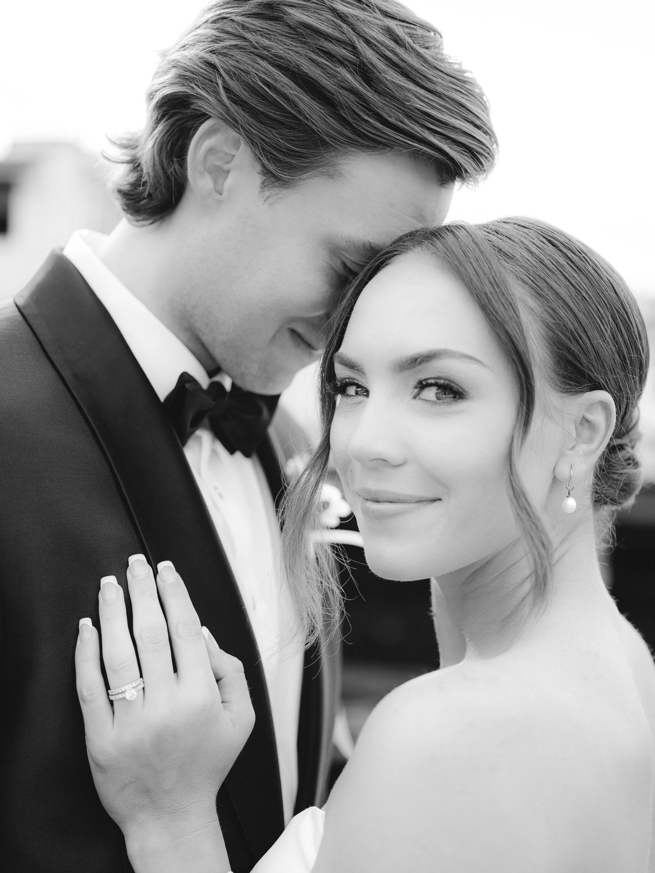 Black-and-white photo of a smiling woman and a man in formal attire, close-up, showing their faces and a hand resting on the man's shoulder.