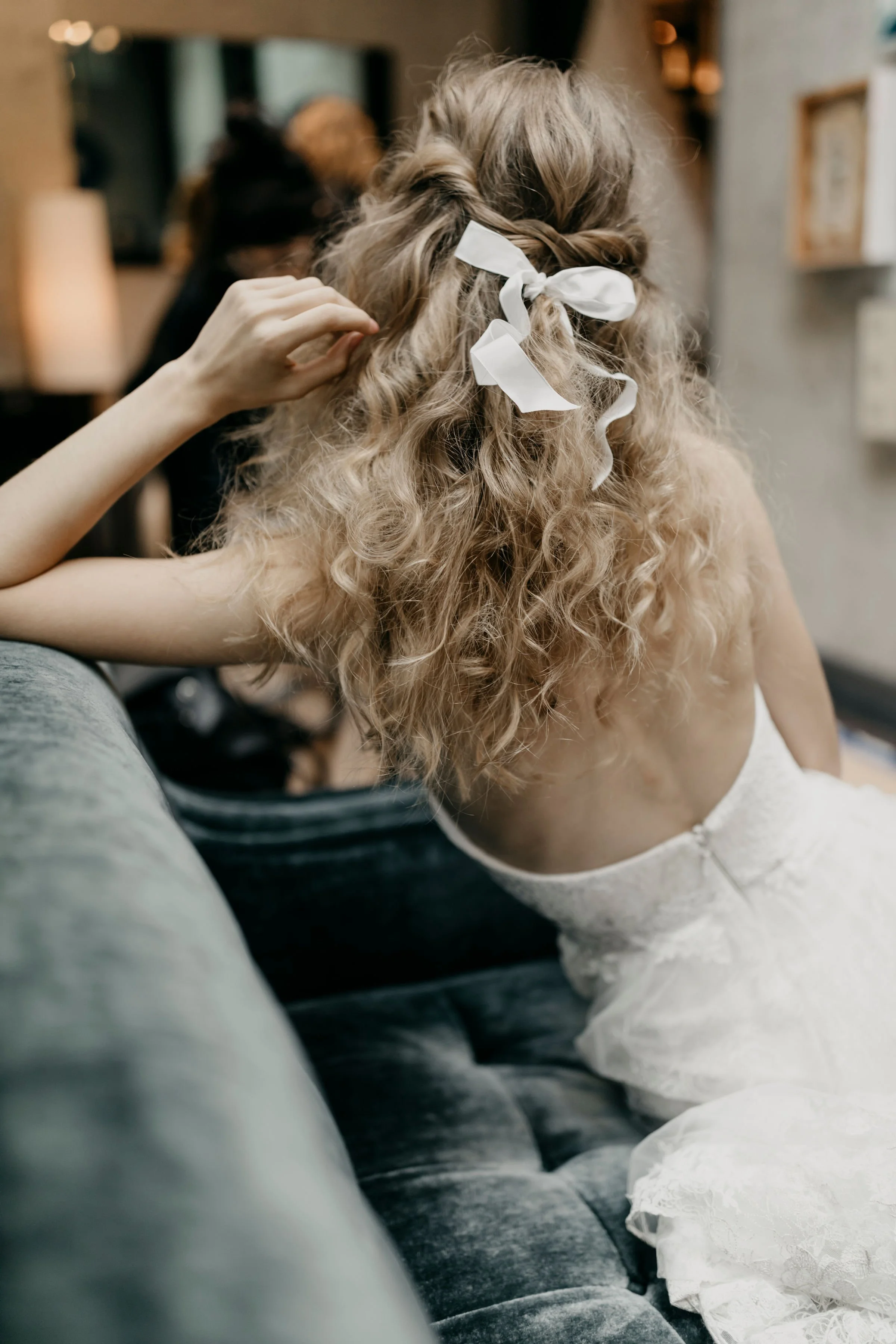 Woman with curly blonde hair, tied with a white ribbon, sitting on a couch, with someone styling her hair in a room with warm lighting.