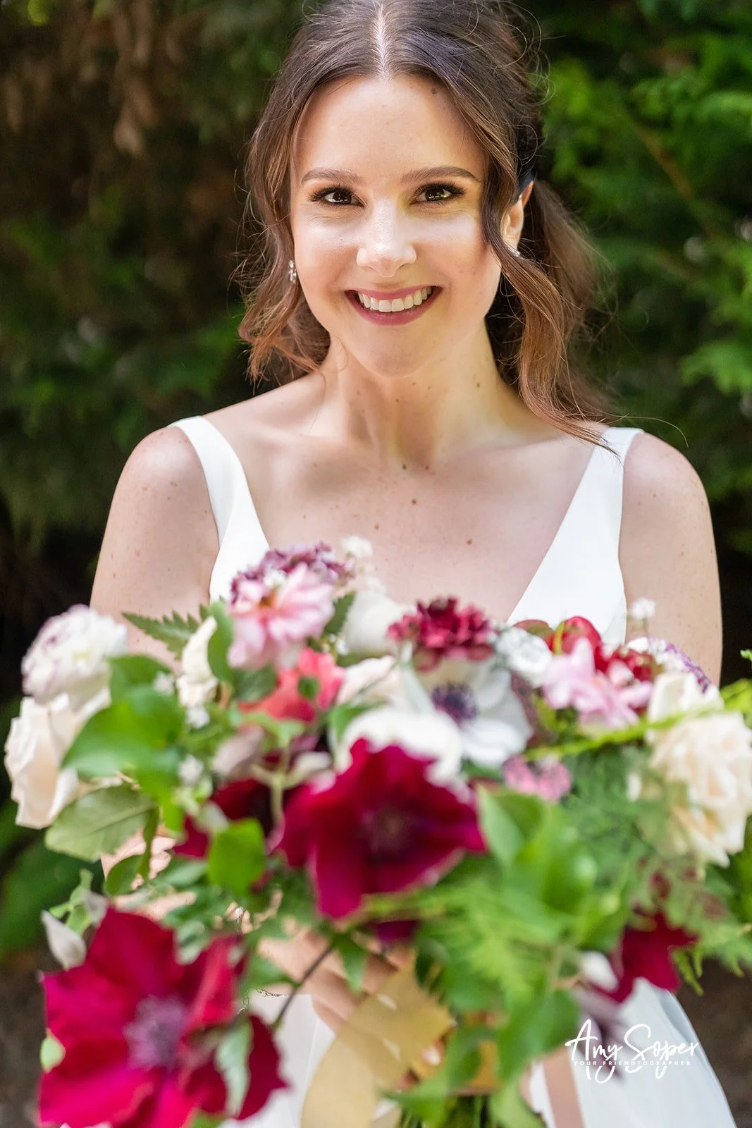 A smiling bride holding a colorful bouquet of flowers outdoors with a green background.