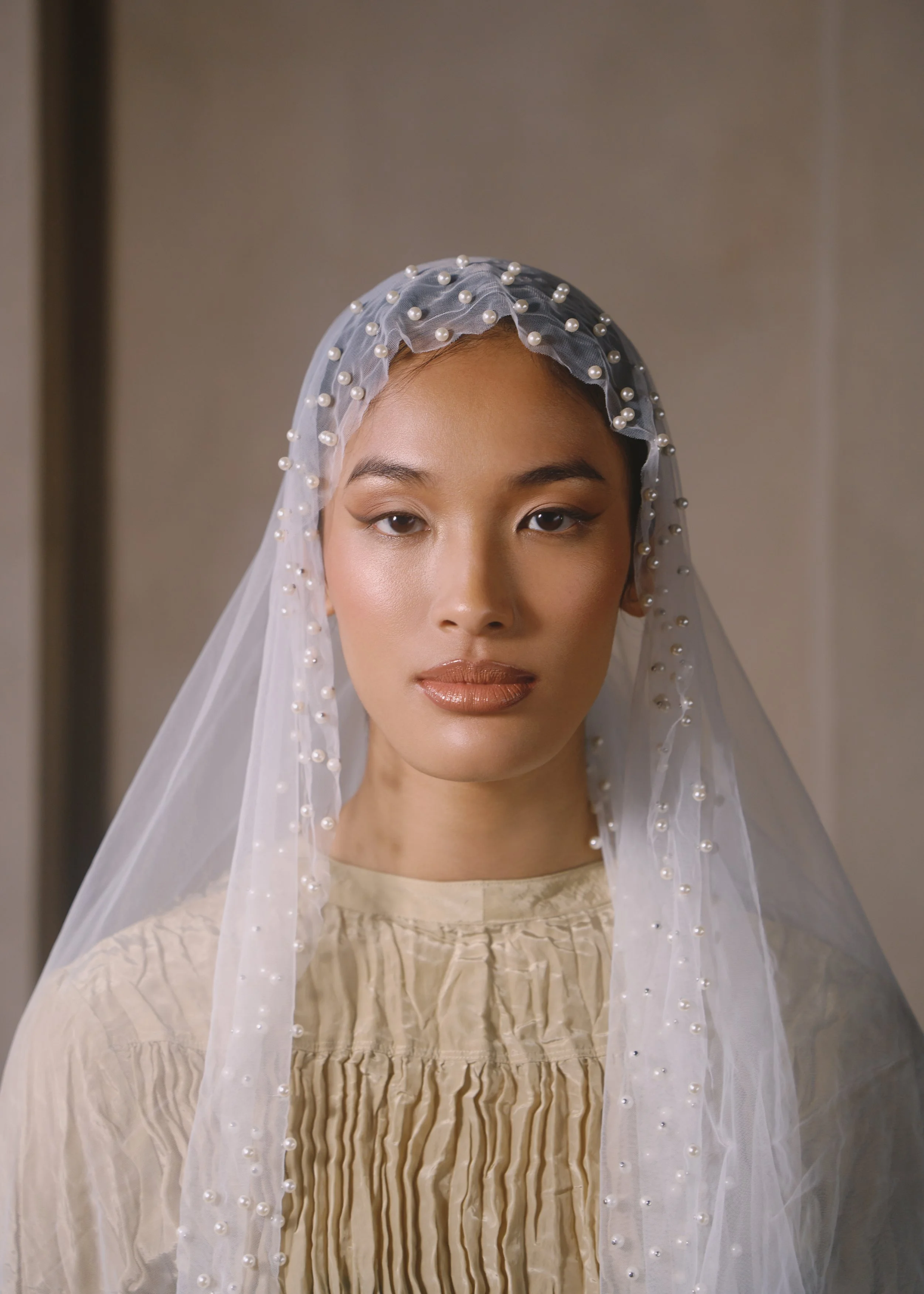 A woman wearing a pearl-embellished veil with a beige dress that has a gathered front detail.