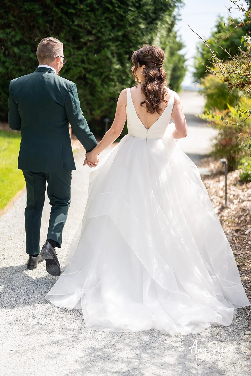 A bride in a white wedding gown holding hands with a groom in a dark suit walking outdoors on a garden path surrounded by green bushes and trees.