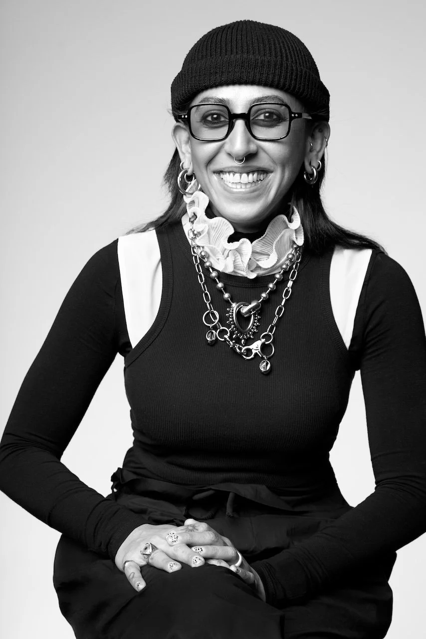 A woman with glasses, a beanie, earrings, layered chains, and a ruffled collar, smiling in a black and white photo.