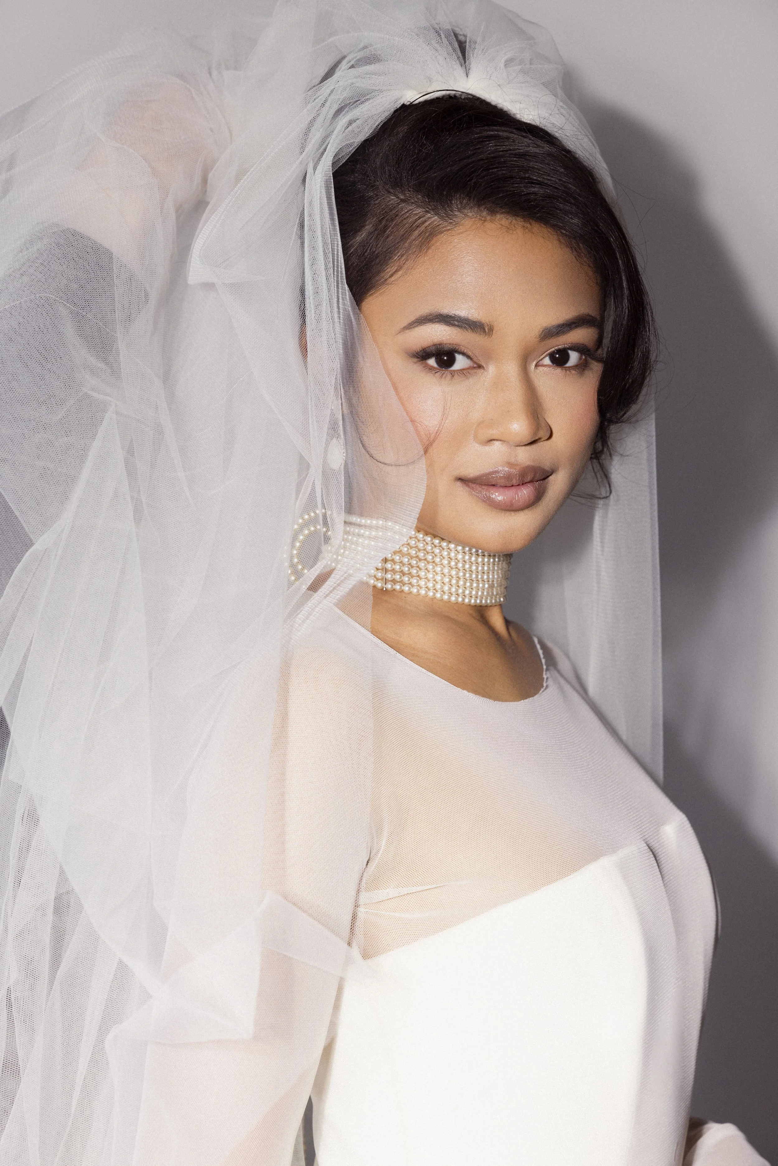 A woman in a white dress with a sheer overlay, wearing a pearl choker necklace, and a white veil, with short dark hair styled to one side, looking at the camera.
Makeup Katya
Hair Meghann
Photographer Andrew Parsons