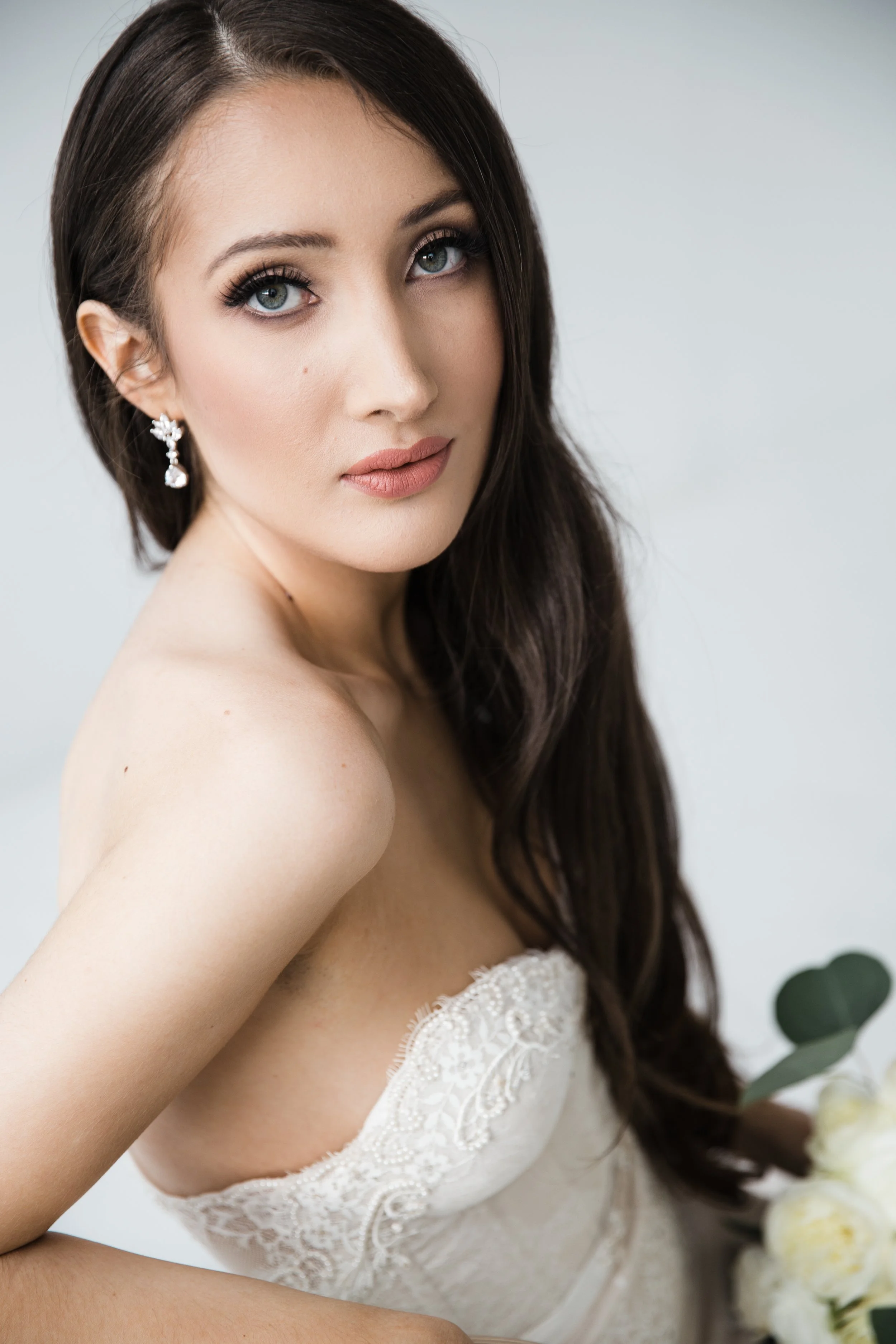 Close-up of a young woman with long dark hair, bright blue eyes, and fair skin, wearing a strapless lace dress and pearl earrings. She is gazing slightly to the side with a neutral expression. There are white roses and green leaves in the lower right