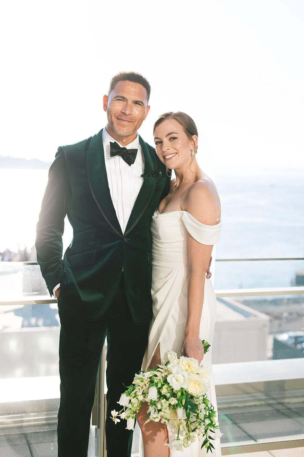A newlywed couple dressed in wedding attire, standing on a balcony with a view of the ocean in the background. The groom is wearing a black tuxedo with a bow tie, and the bride is in a white off-shoulder wedding dress holding a bouquet of white flowe