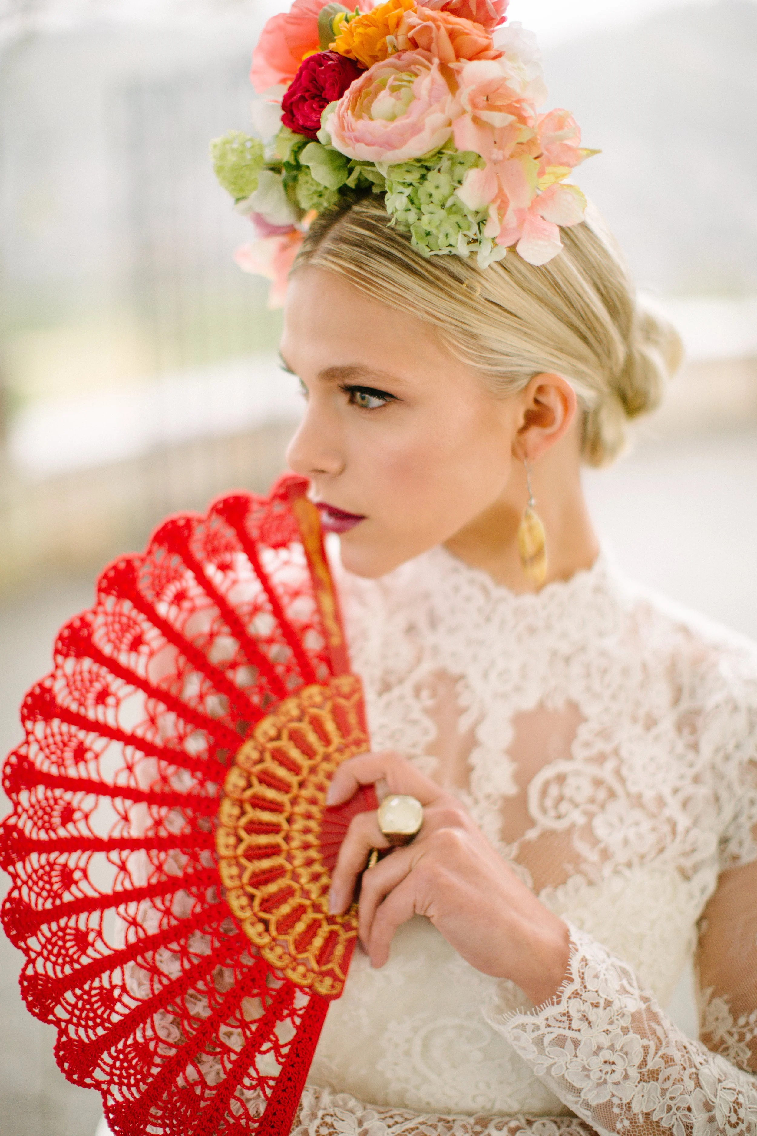 A woman with blonde hair in an elegant updo, wearing a white lace dress, adorned with a large colorful floral headpiece, holding a red lace fan, and wearing a large ring and drop earrings.