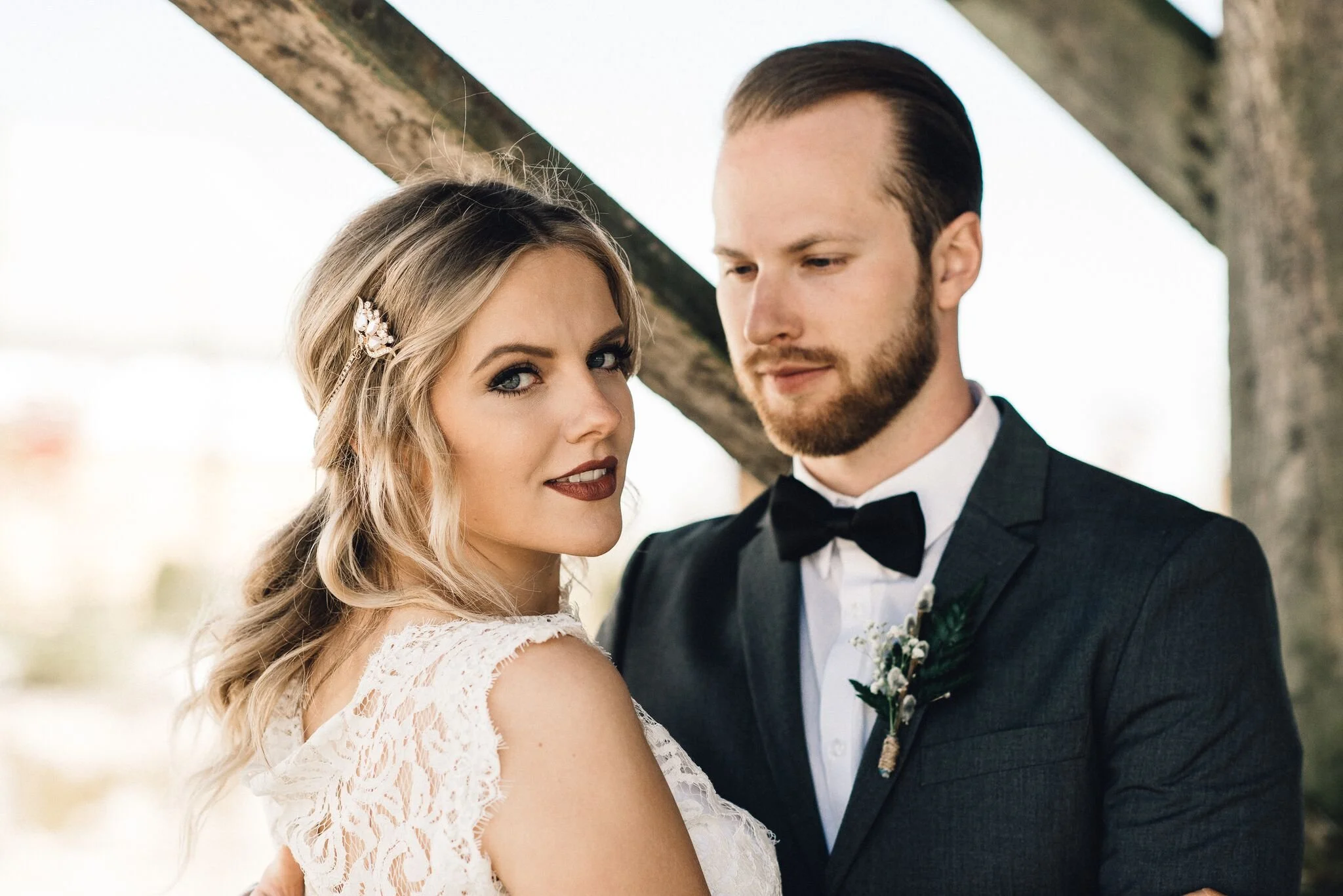 Bride and groom posing outdoors during their wedding, leaning against a wooden beam, with the bride looking at the camera and the groom looking slightly away.