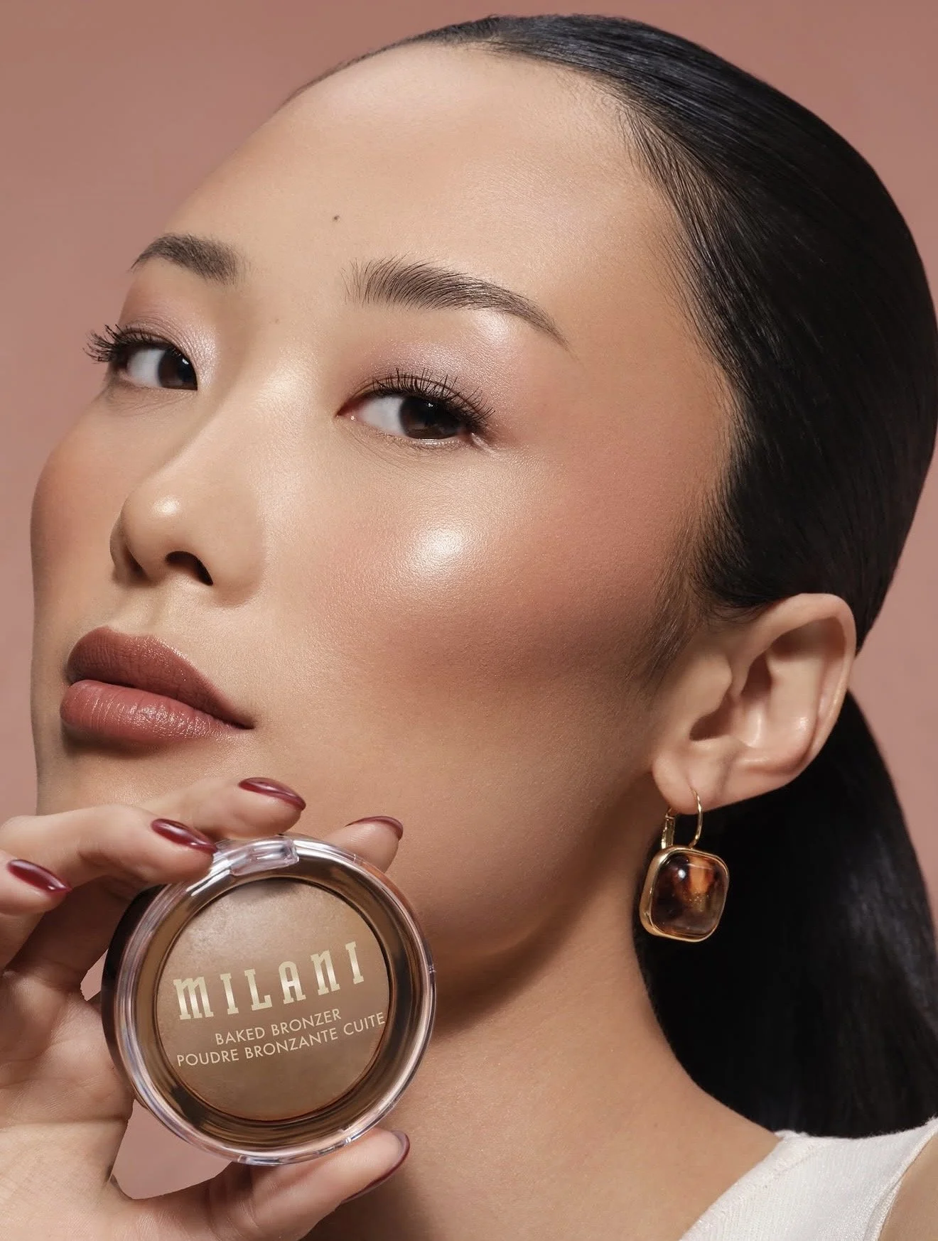 Close-up of a woman with flawless skin, dark hair slicked back, and wearing a gold earring with a large tortoise shell pendant. She holds a compact labeled 'Milani Baked Bronzer.'