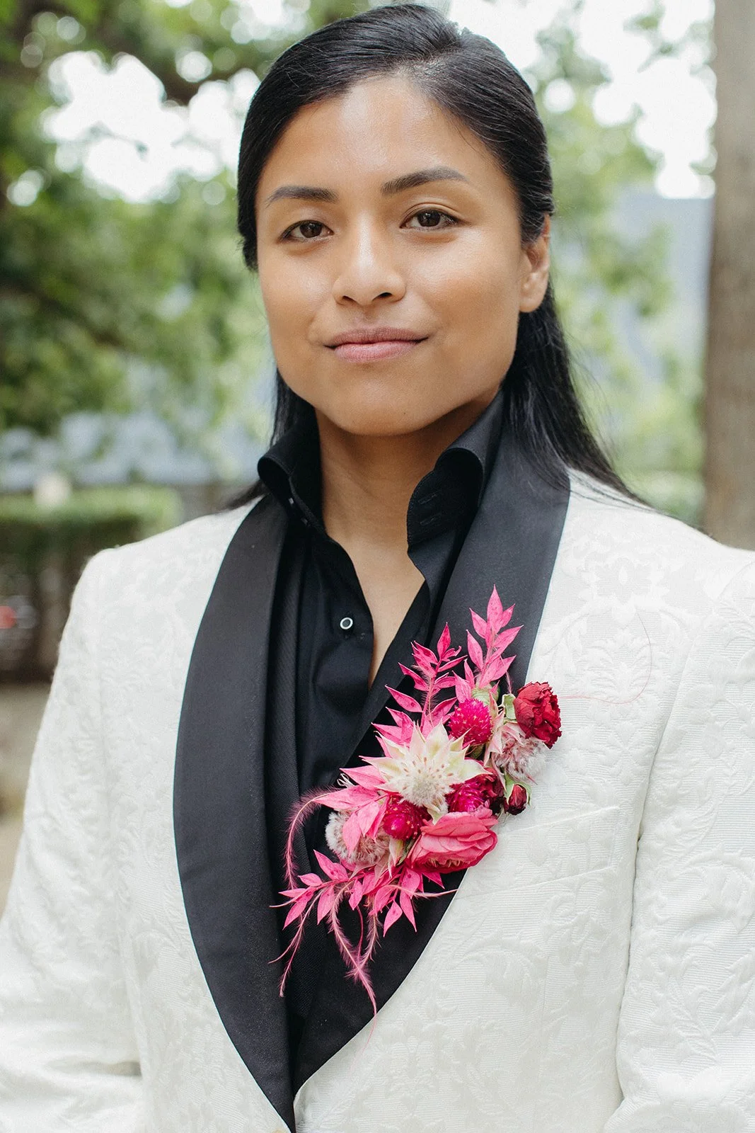 A woman with black hair styled back, wearing a white blazer with floral embroidery, a black shirt, and a pink floral corsage, standing outdoors with blurred trees in the background.