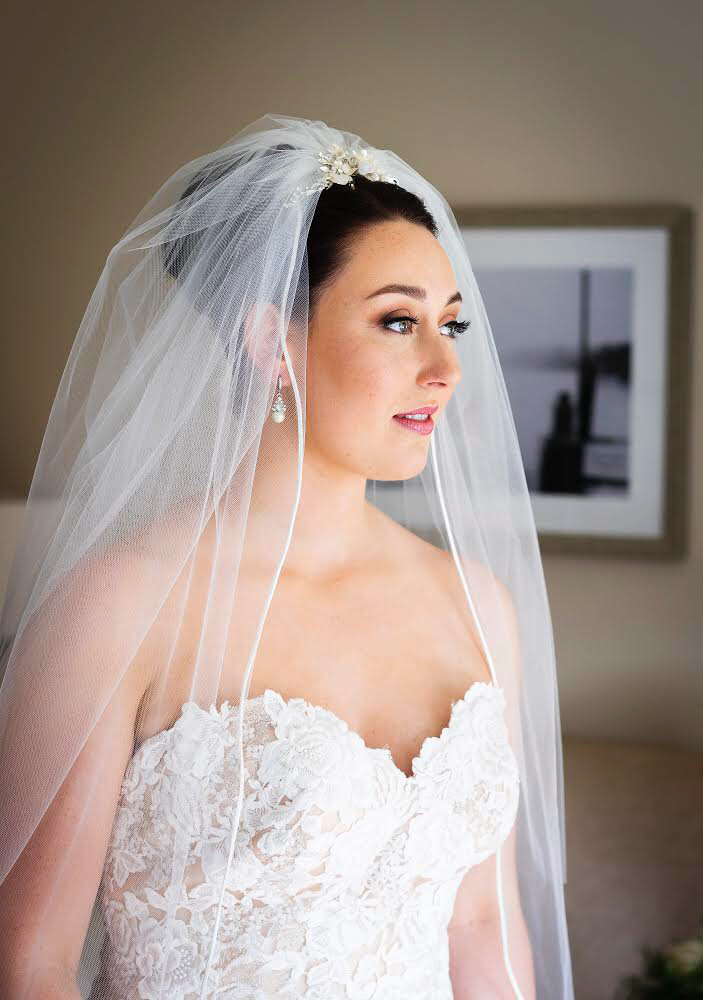 A bride wearing a lace wedding dress and veil, with pearl earrings and a floral hairpiece, looking to the side indoors.
