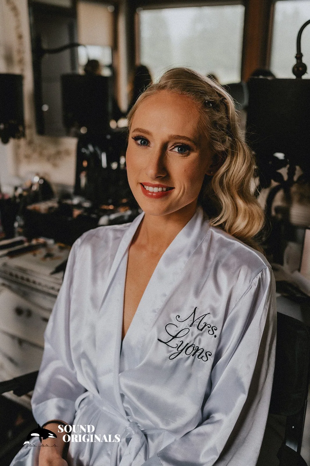 A woman with blonde hair styled in loose waves, wearing a white satin robe with 'Mrs. Lyons' embroidered on it, smiling in a makeup room with various makeup and hair styling tools in the background.