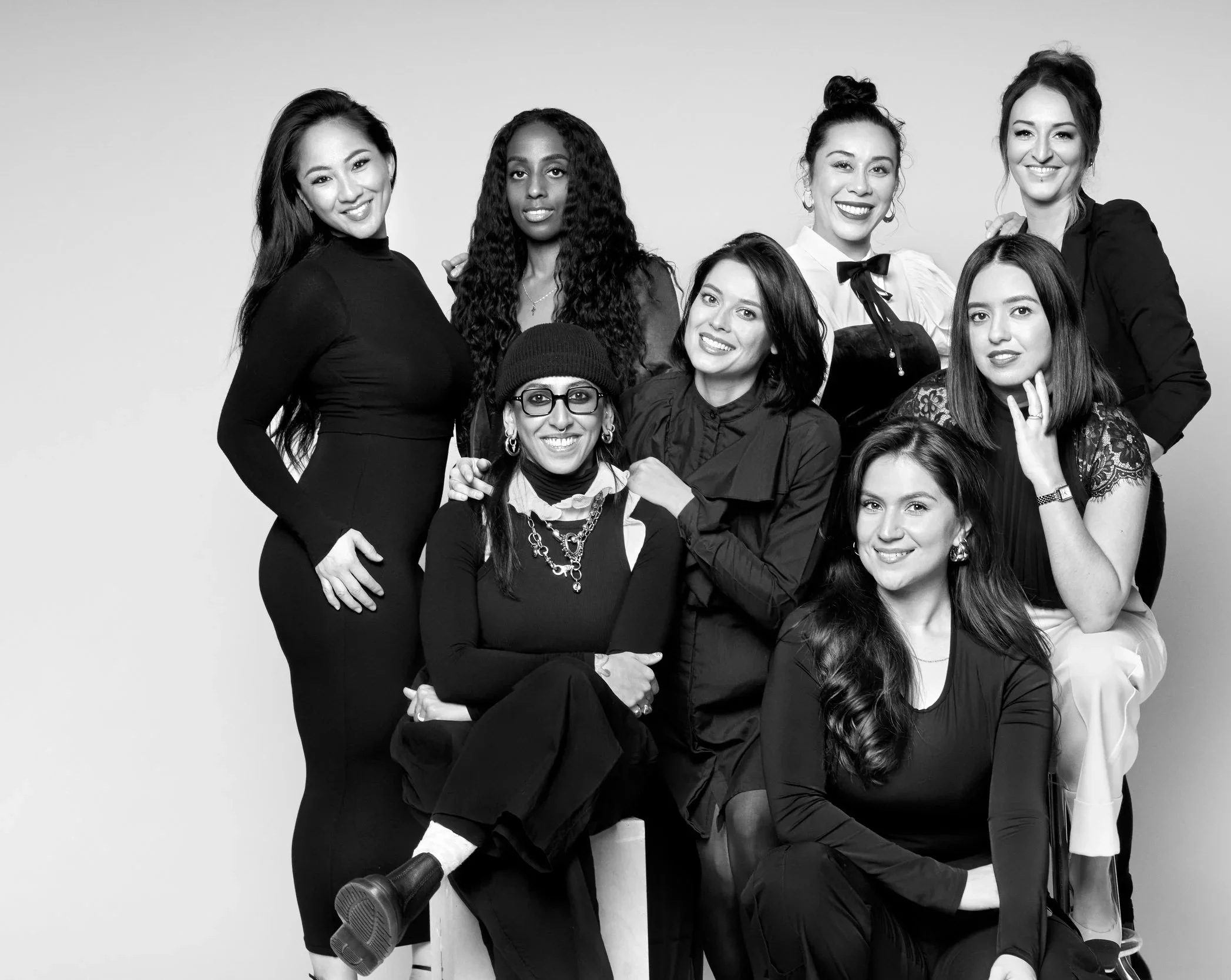 A group of diverse women smiling, posing together against a plain background. They are dressed in stylish clothing, with some wearing accessories like glasses and jewelry.