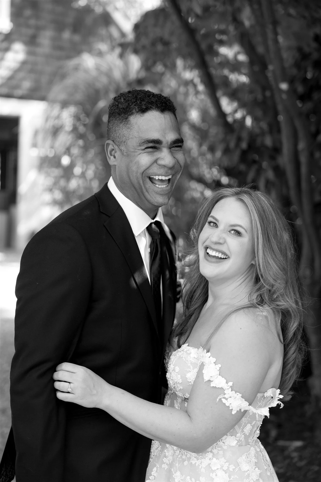 A smiling man in a tuxedo and a smiling woman in a lace wedding dress posing outdoors. They are embracing each other, with the man’s arm around the woman’s waist and her hand on his chest, showing her wedding ring. The background features blurred tre