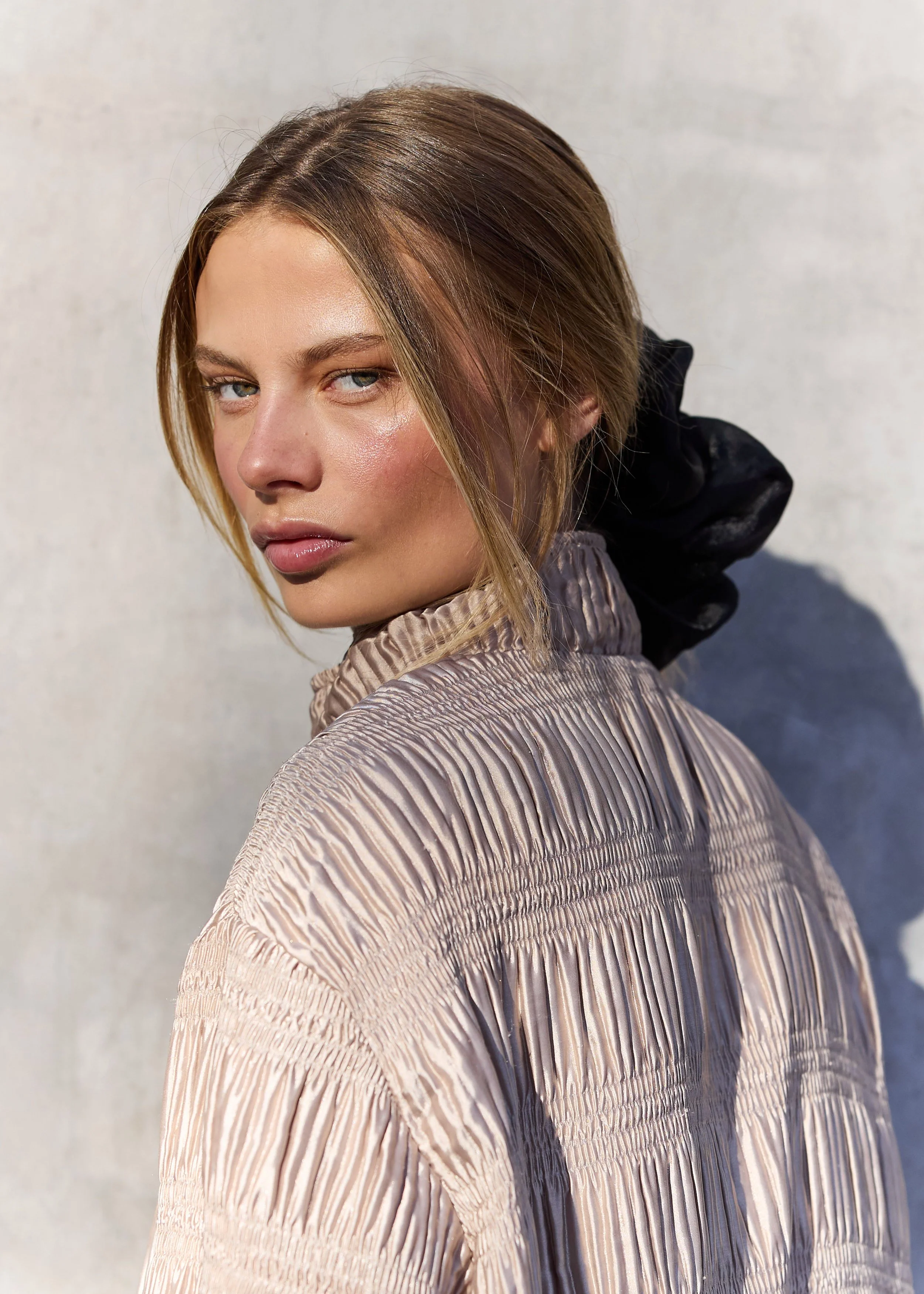 Close-up of a woman with short, light brown hair, wearing a textured light pink blouse, leaning slightly forward with a neutral expression against a plain background.