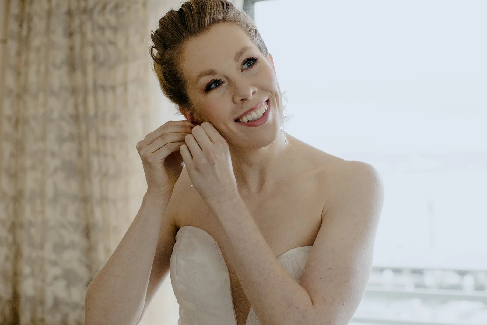 A woman with red hair and blue eyes smiling as she puts on earrings, wearing a strapless white dress.
