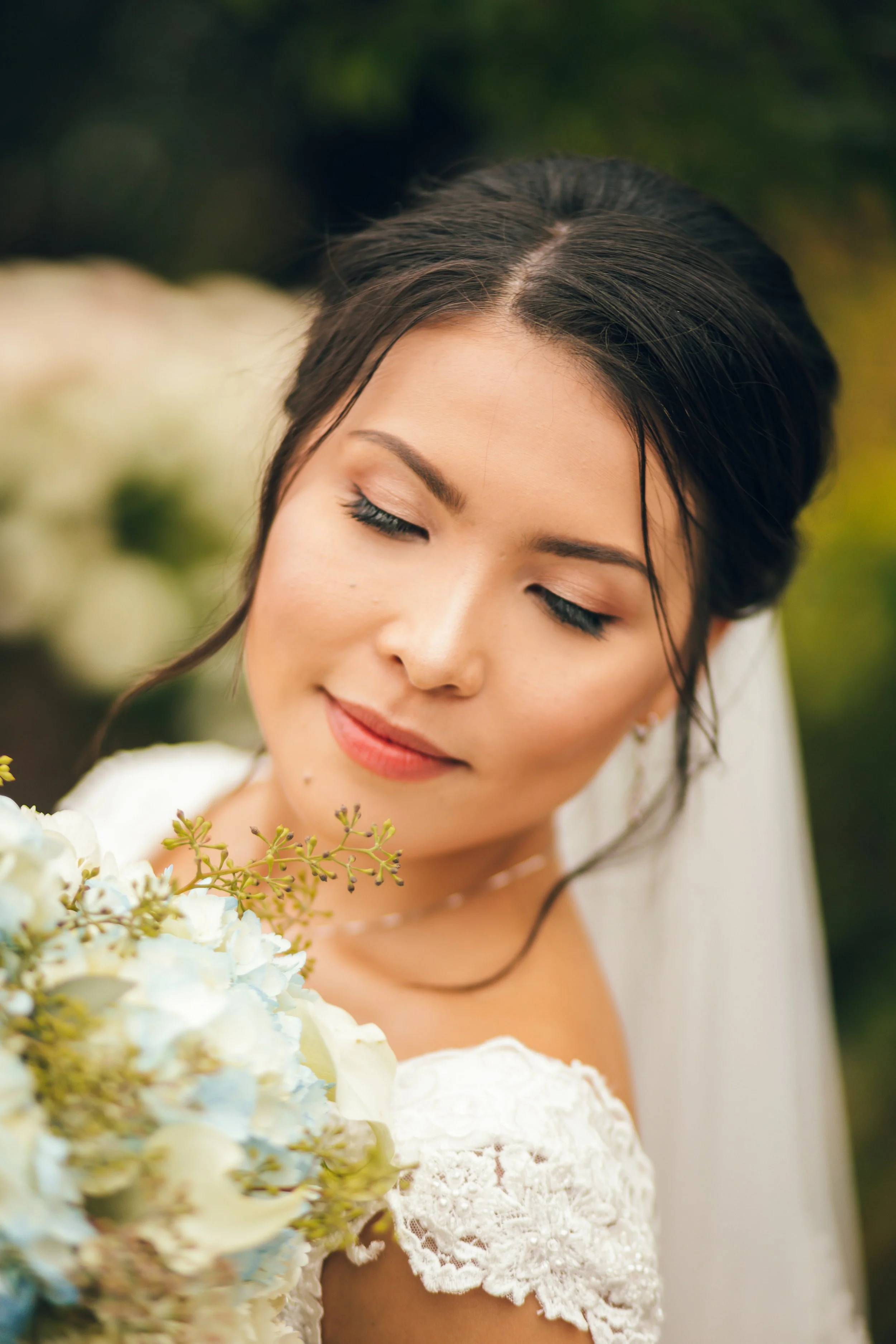 A close-up of a woman with dark hair styled neatly, eyes closed, on a sunny outdoors background, smiling softly, dressed in a white lace dress, holding a bouquet of white and light blue flowers.