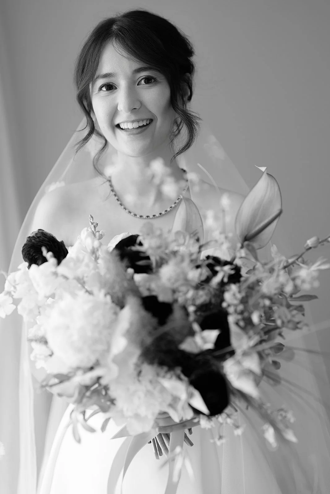 Black and white photo of a smiling woman with short hair, wearing a pearl necklace, holding a large bouquet of flowers, and wearing a wedding dress with a veil.